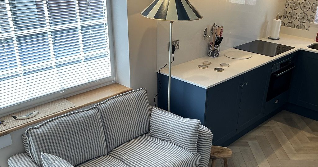 Small striped sofa near a window with blinds, beside a modern kitchen counter and floor lamp