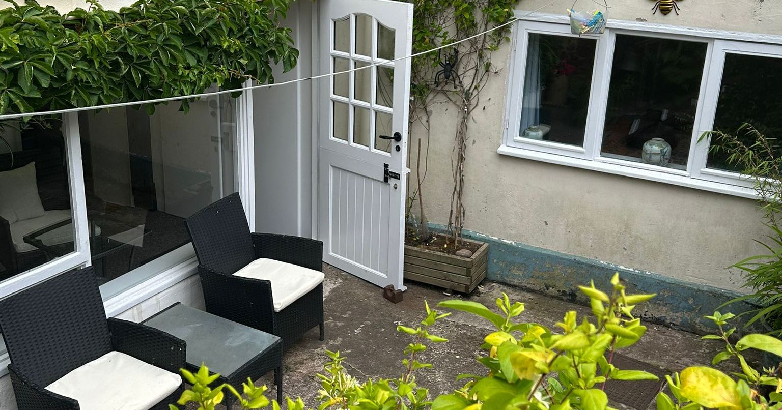 Outdoor courtyard with two black chairs and white cushions, a small table, green plants, white door, wall-mounted bee decorations, and window