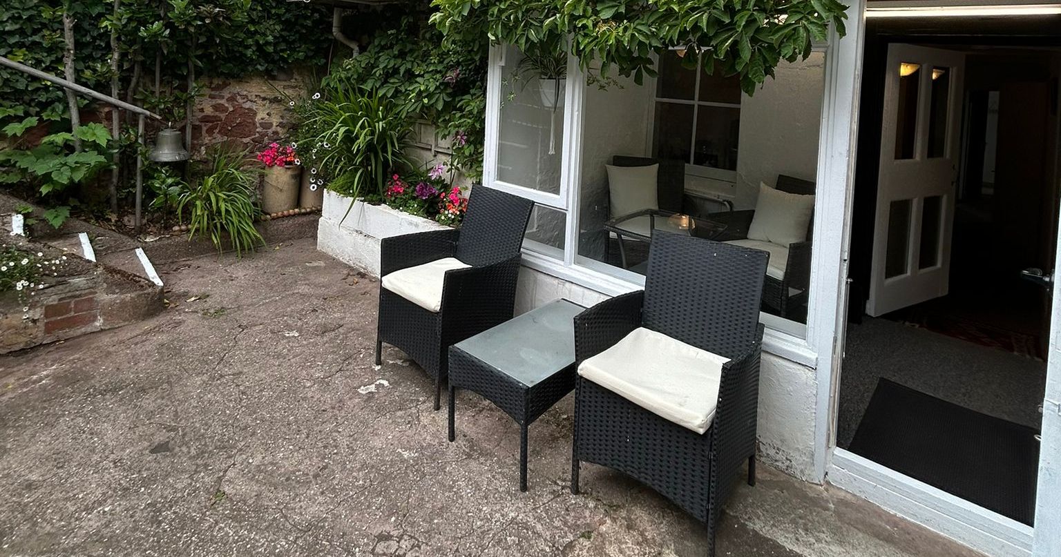 Outdoor terrace area with two chairs, a small table, and garden plants in front of a house with large windows.