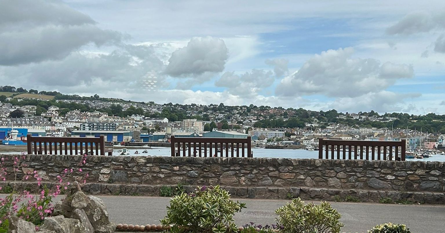A scenic view of a waterfront town with benches, stone wall, garden, and cloudy sky.