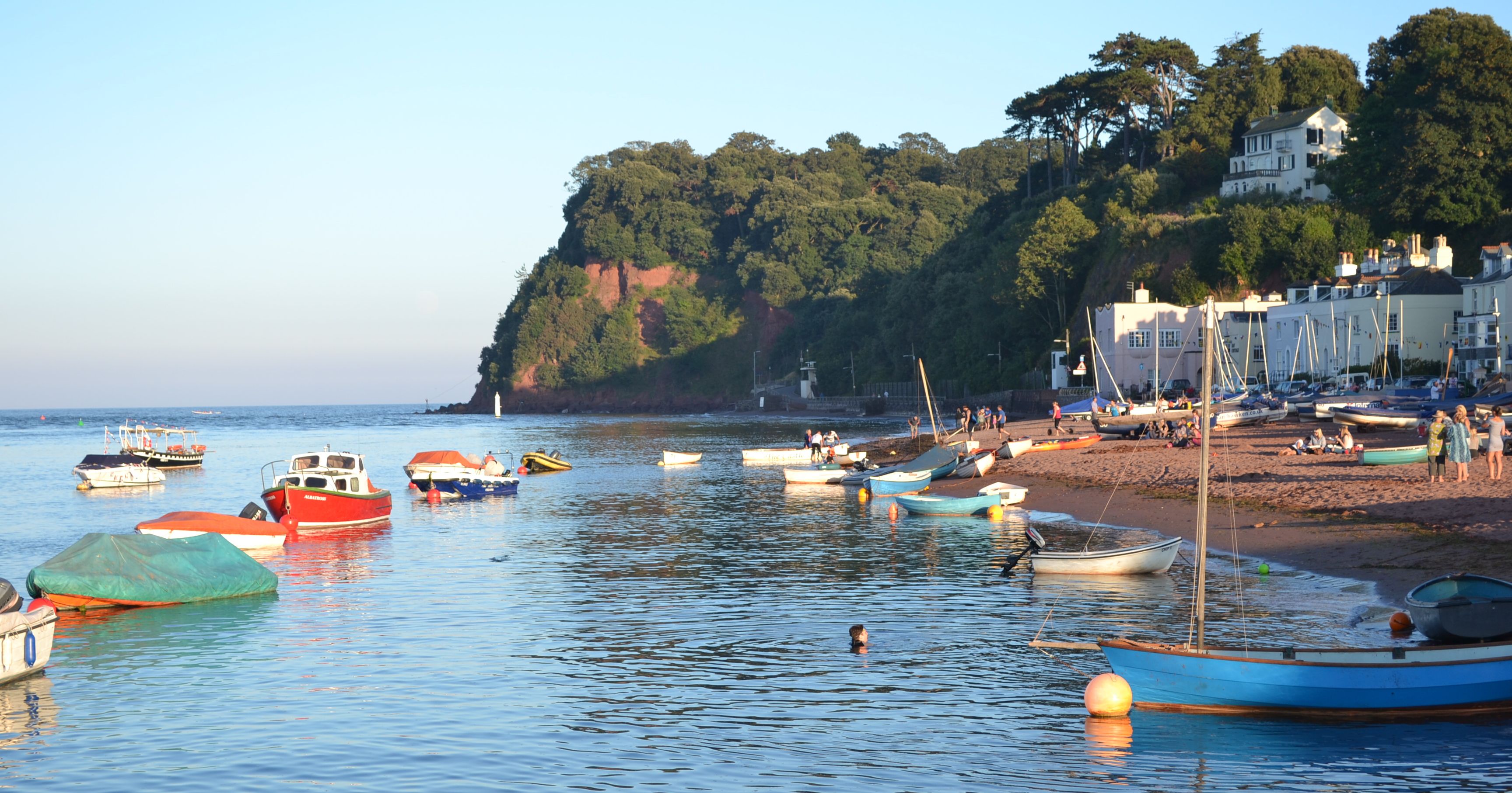 A calm seaside scene with small boats anchored near the shore, people on the sandy beach, and houses surrounded by trees on a hillside.