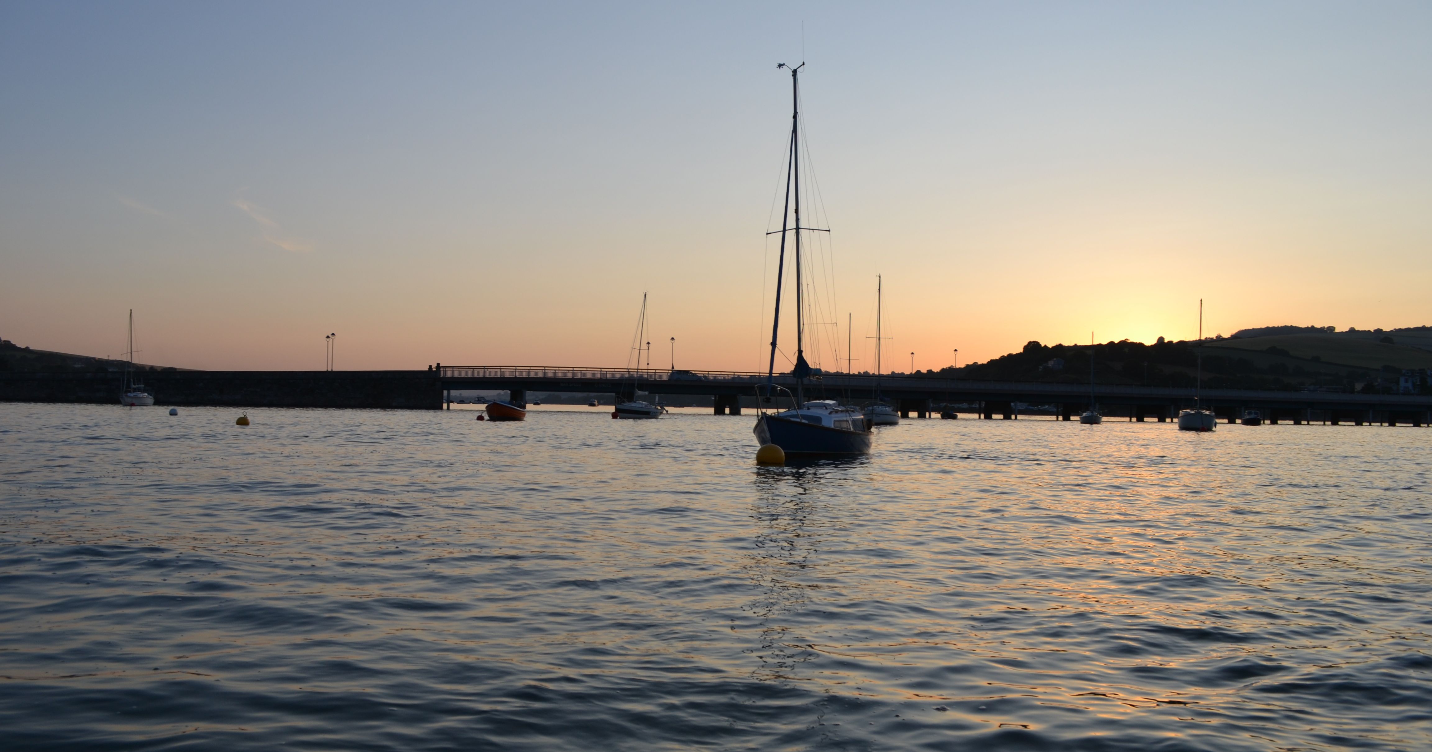 Sailboats on calm water at sunset with a bridge and hills in the background.