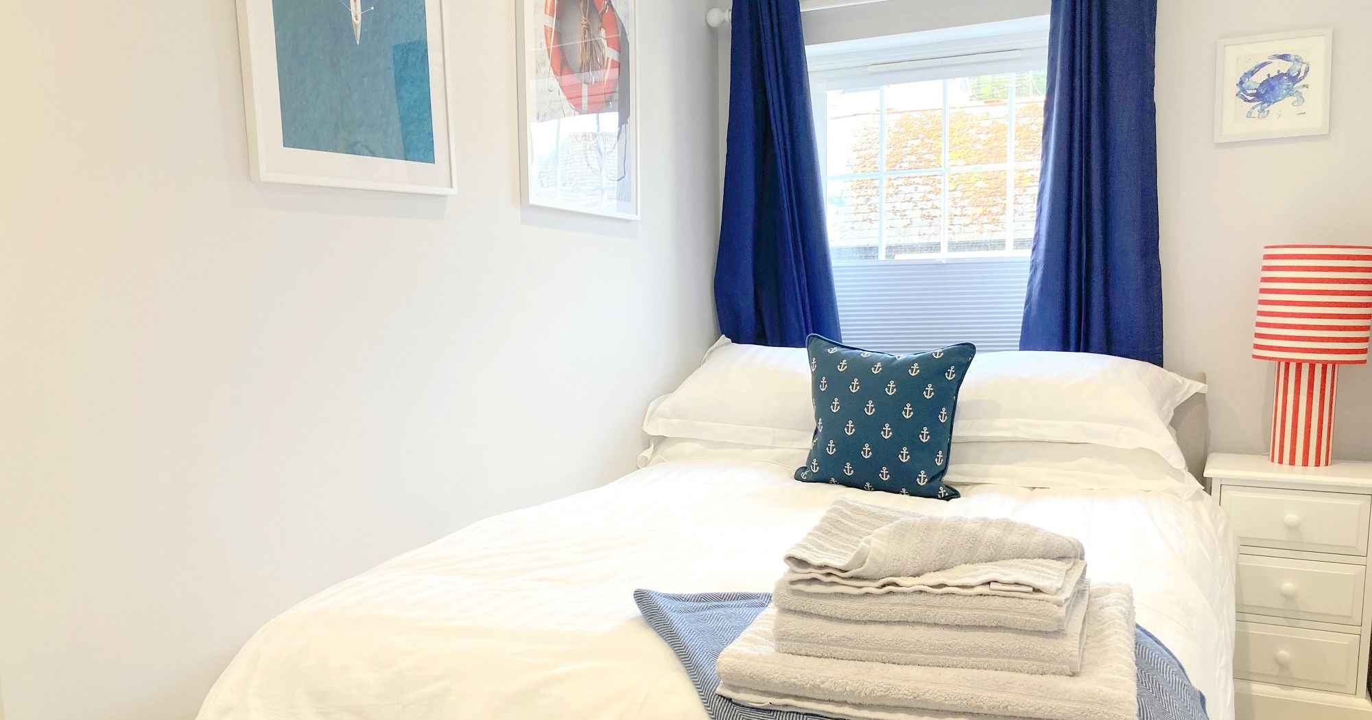 Cozy bedroom with nautical decor featuring a double bed, blue curtains, striped stool, and folded towels on the bed.