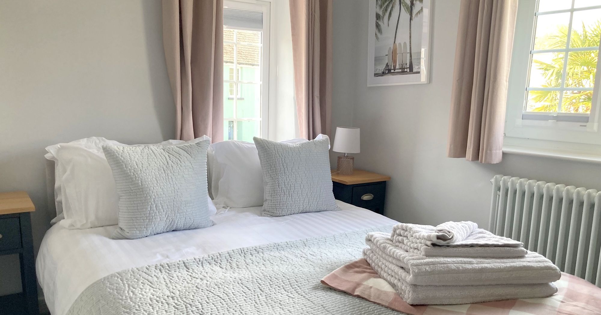 Bright bedroom with a king-sized bed, soft grey bedding, stacked white towels, and a window letting in natural light.