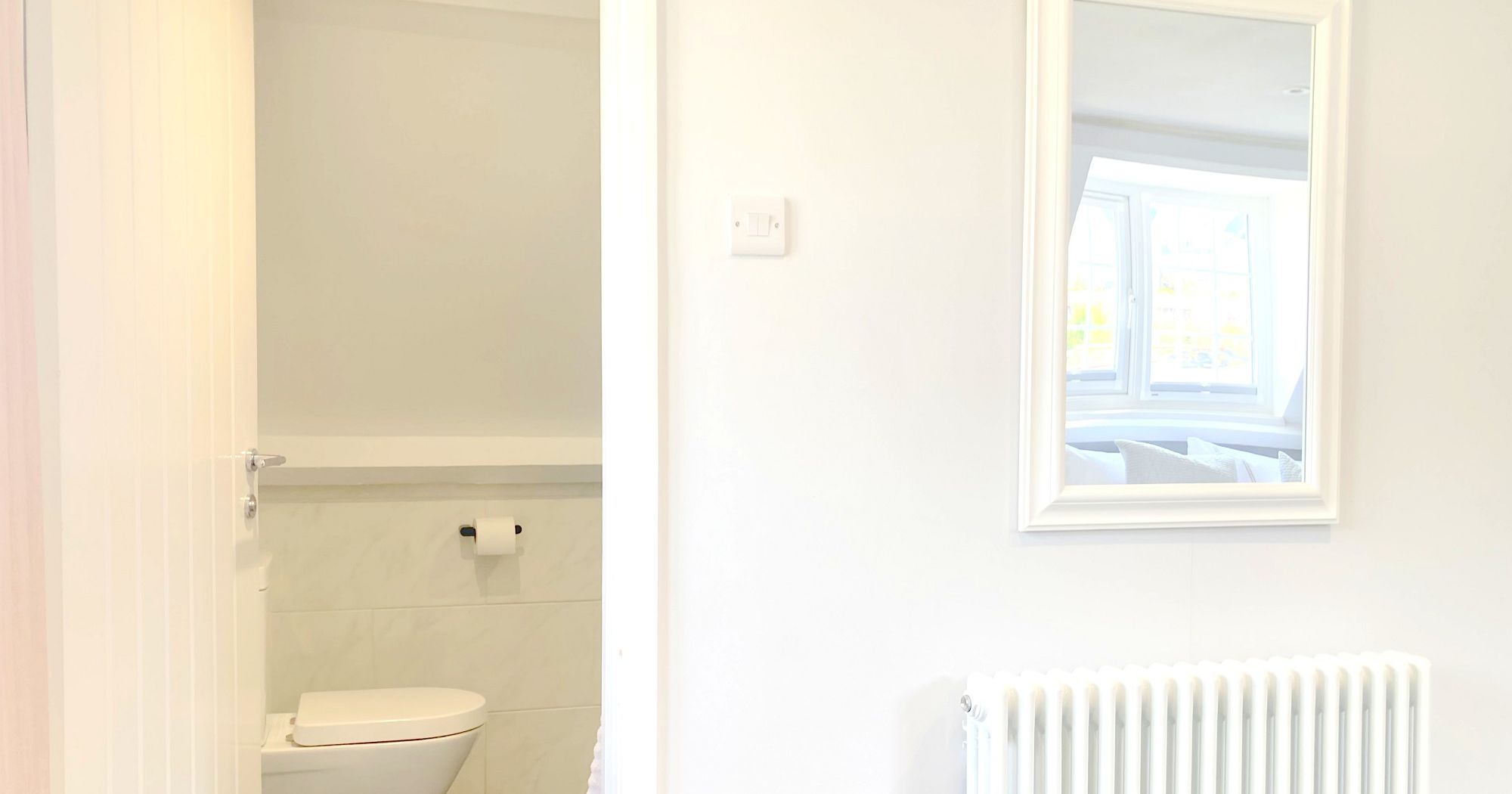 Modern bathroom with an open door revealing a toilet and a wall-mounted toilet paper holder, with a white radiator and framed mirror on the adjacent wall.