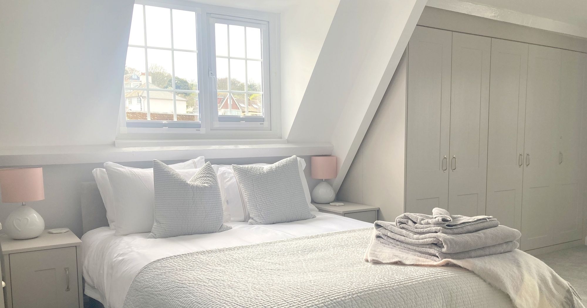 Bright master bedroom with king size bed, white bedding, grey accent pillows, neatly folded towels, and built-in wardrobes.