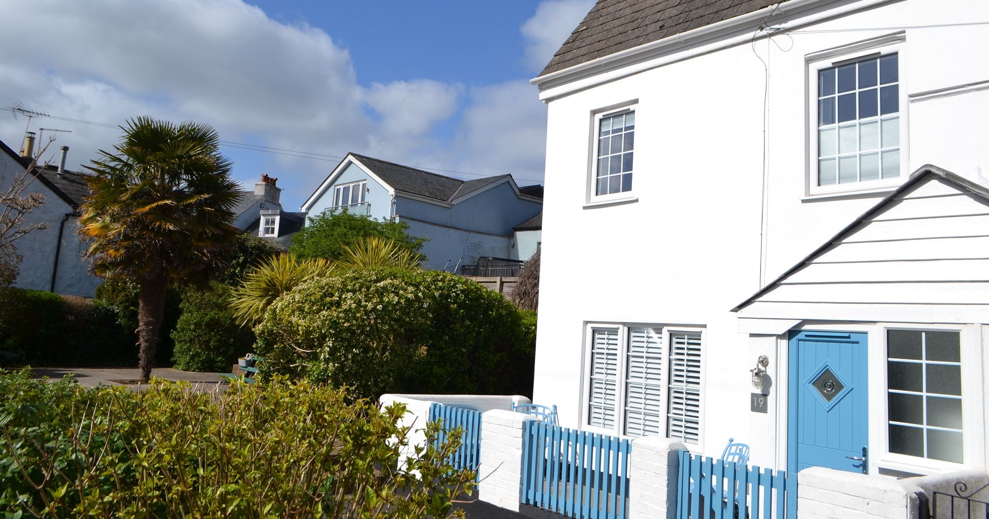 White cottage with blue door and picket fence beside palm trees and bushes under a blue sky.