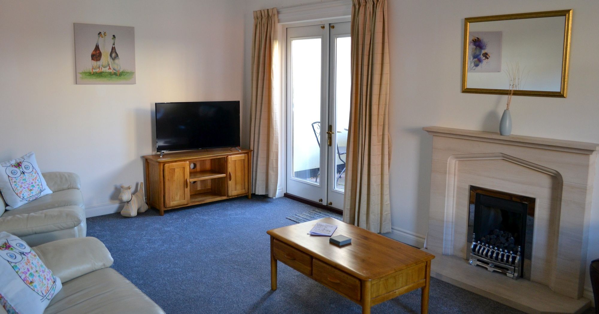Living room with beige sofas, wooden coffee table, TV on wooden stand, fireplace, and French doors with curtains.