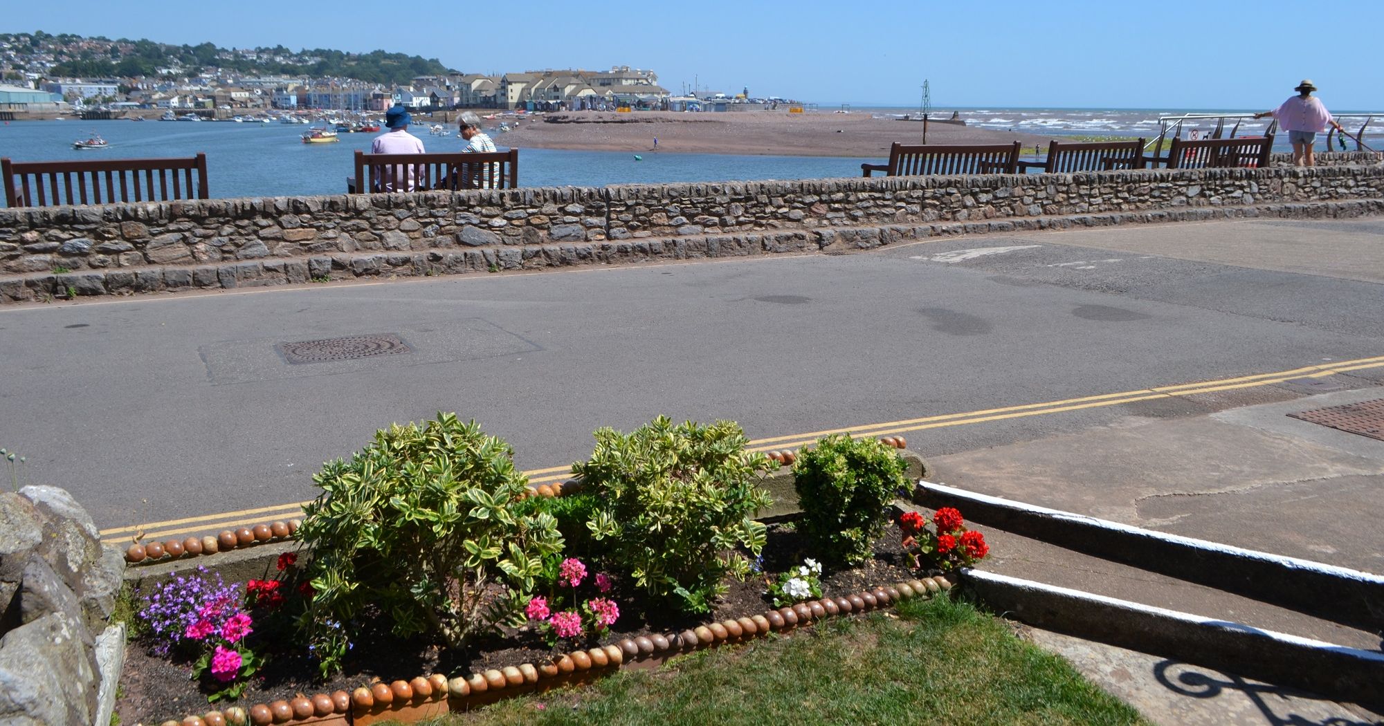 Seaside promenade with benches overlooking the sea, a stone wall, and a flower bed in the foreground.