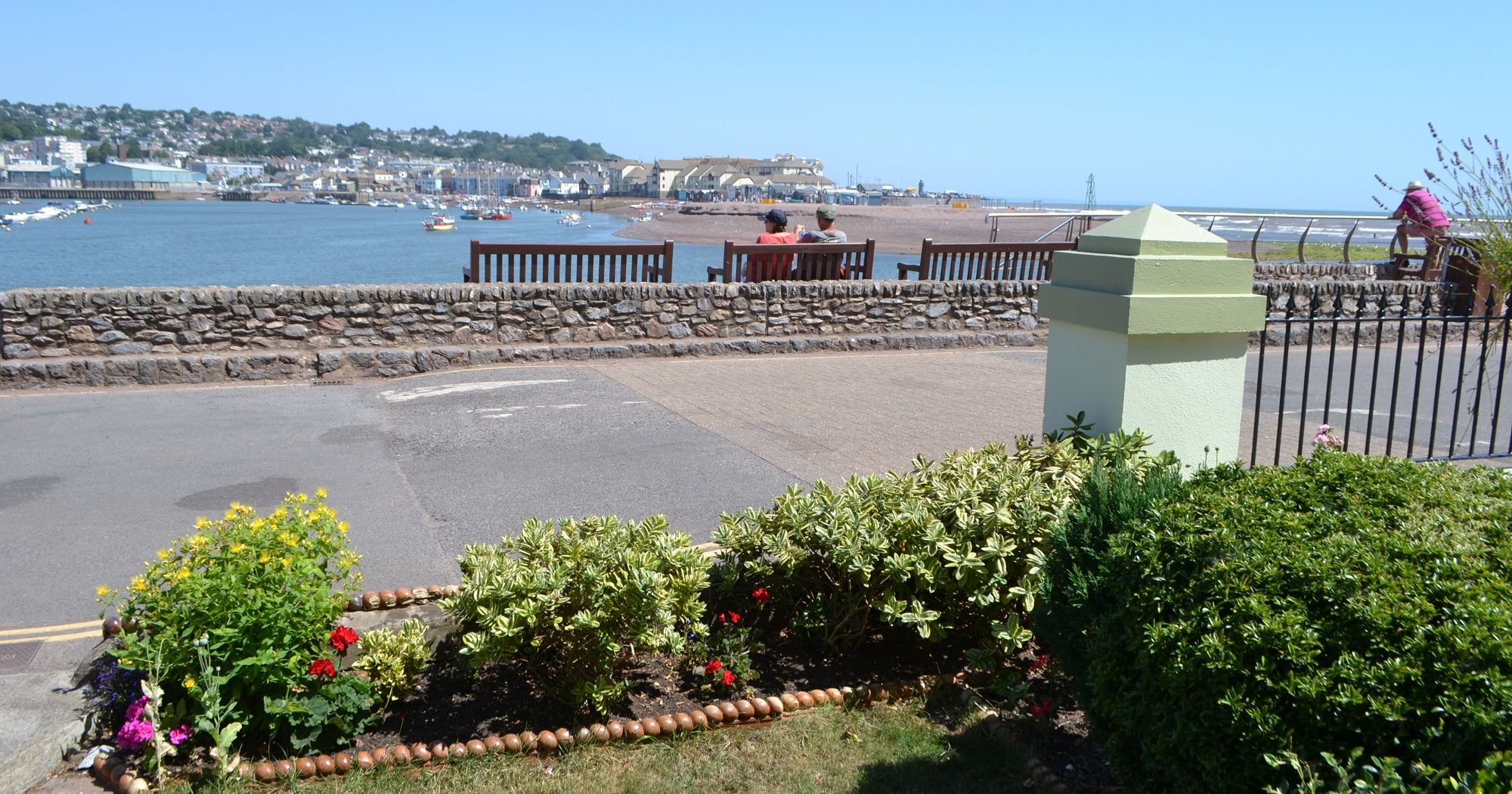 A view of a seaside town with benches facing the water, people sitting and relaxing, a garden in the foreground, and boats anchored in the harbor.