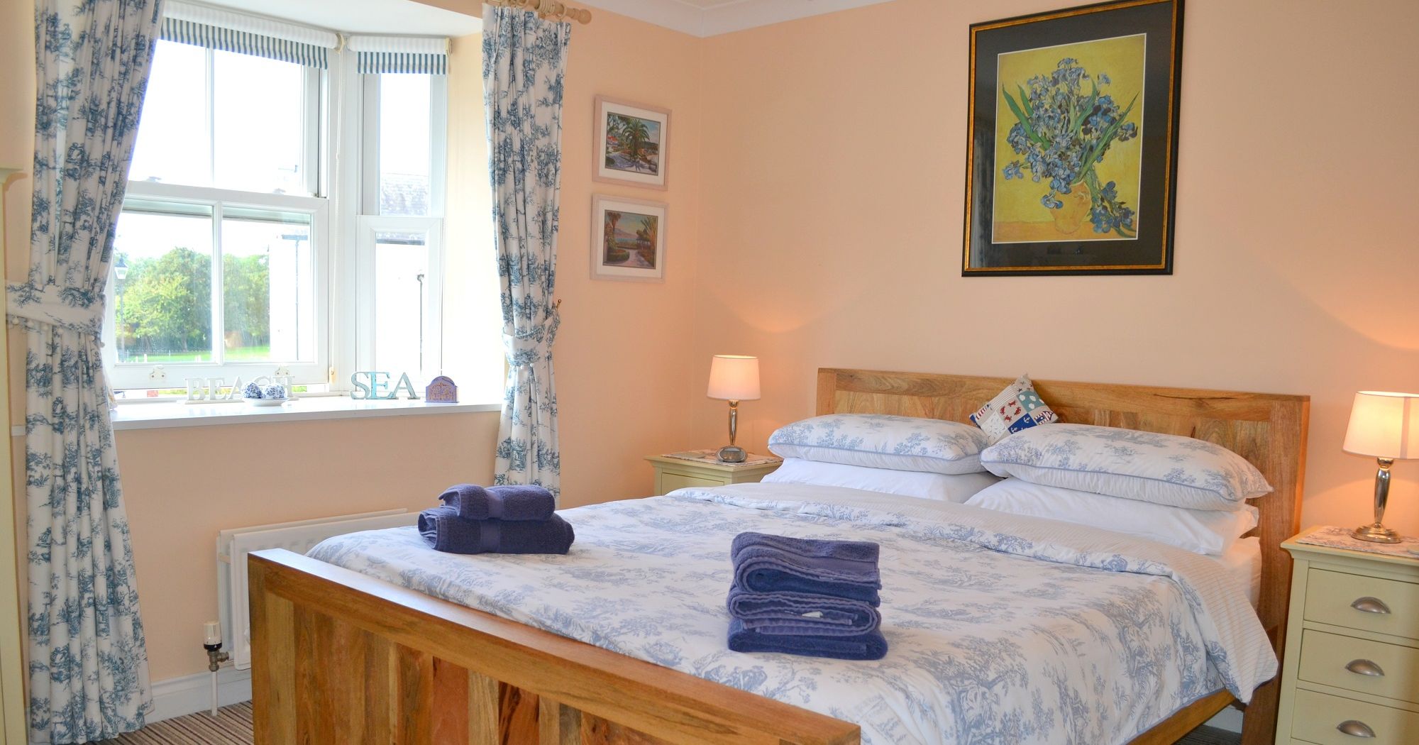 Bright and cozy bedroom with large wooden bed, blue and white bedding, bedside tables with lamps, and a window with patterned curtains.