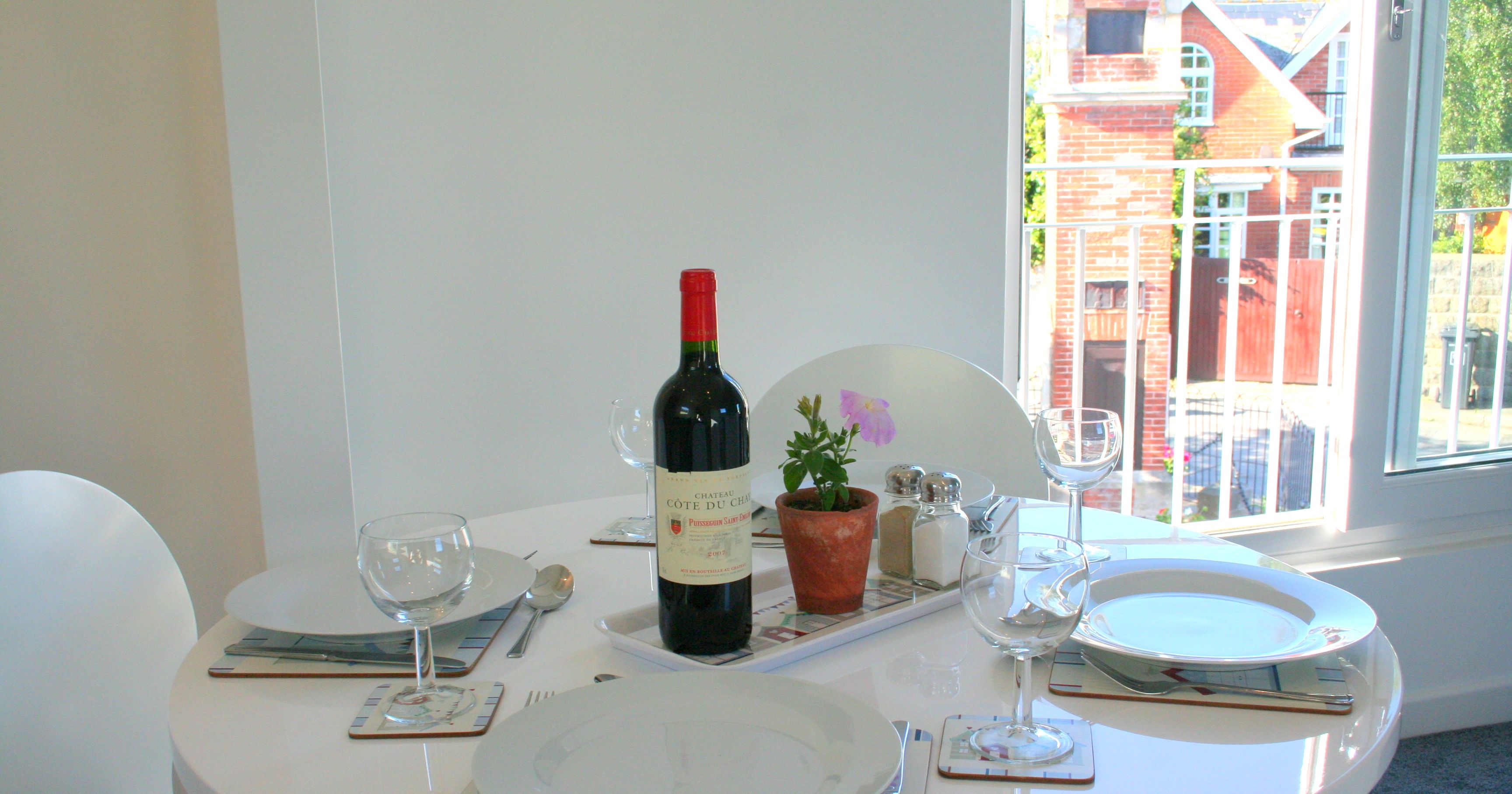 Dining table set for three with wine bottle, flower pot, and a window view showing a brick clock tower