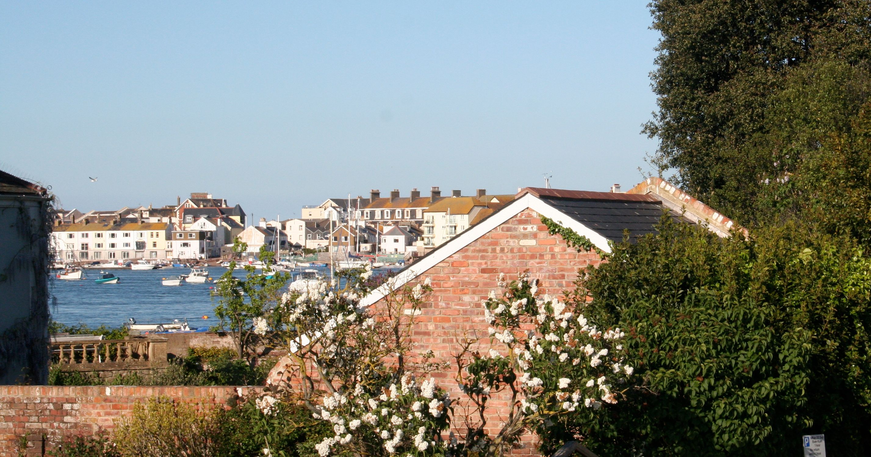 View of a waterfront village with boats on the water, a brick cottage, flowering trees, and greenery in the foreground under a clear blue sky.