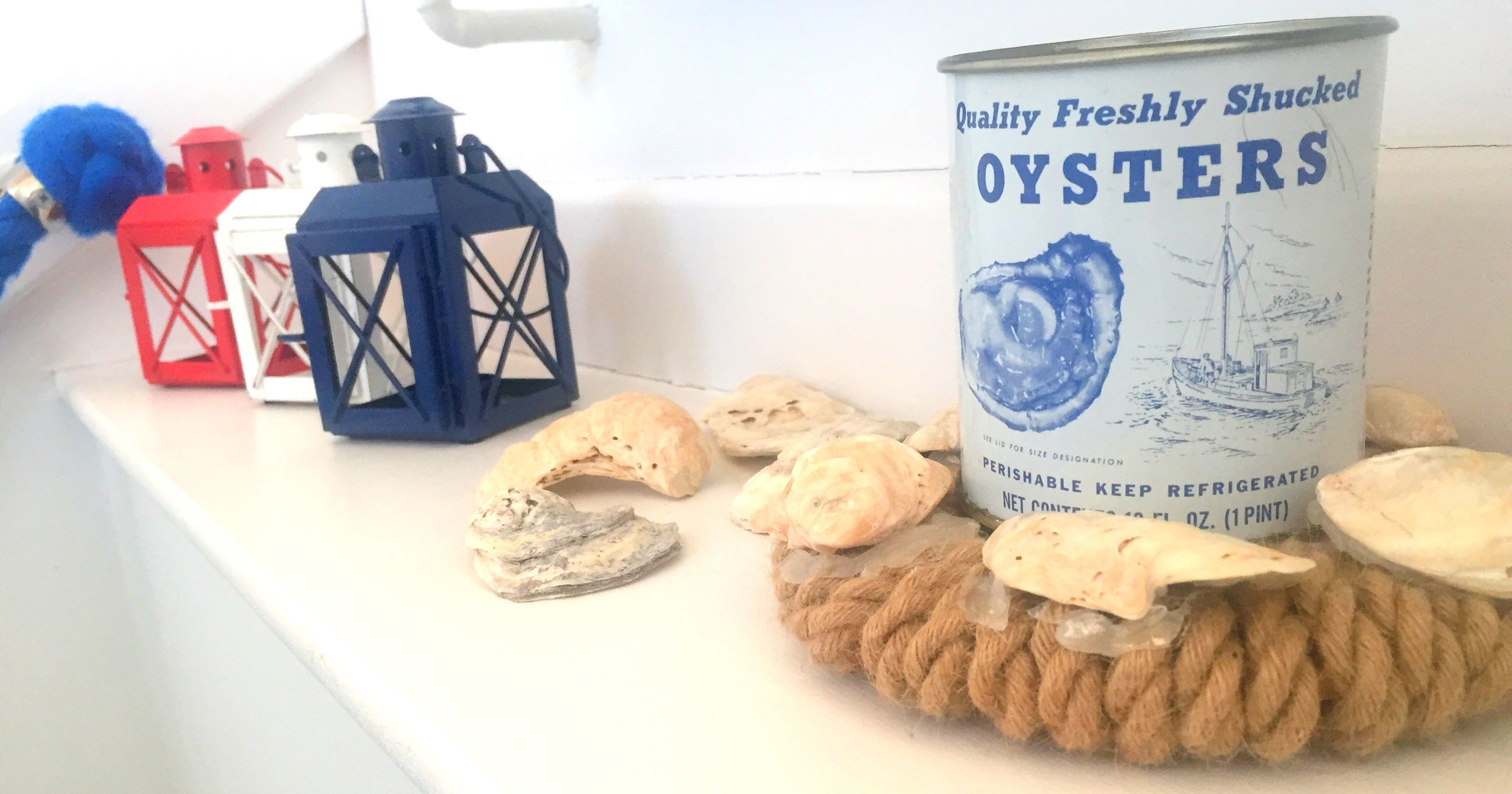 A can of oysters surrounded by seashells and nautical-themed lanterns on a white shelf.