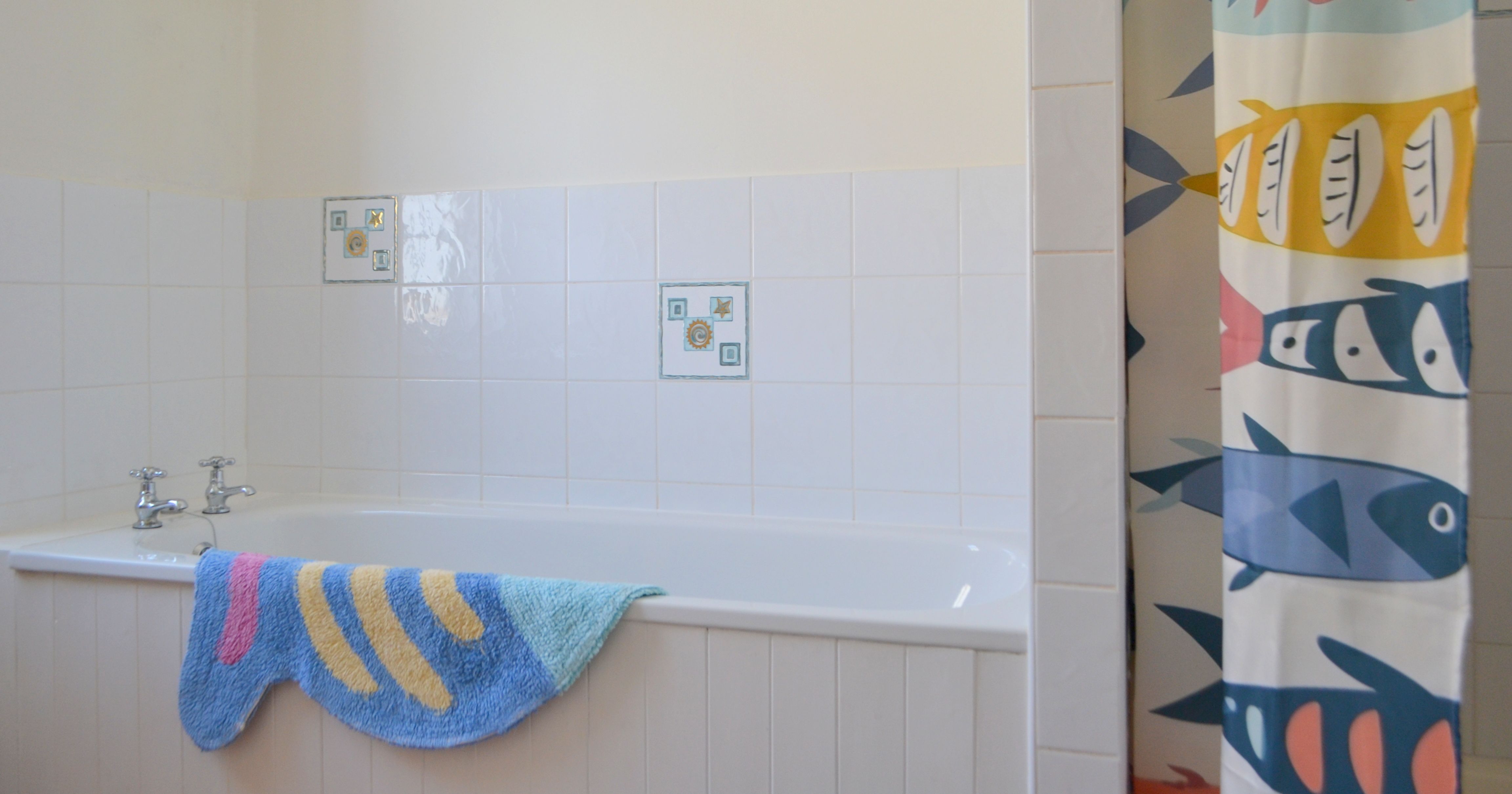 Bright bathroom with a white tub, blue and yellow bath mat, and colorful fish-patterned shower curtain.