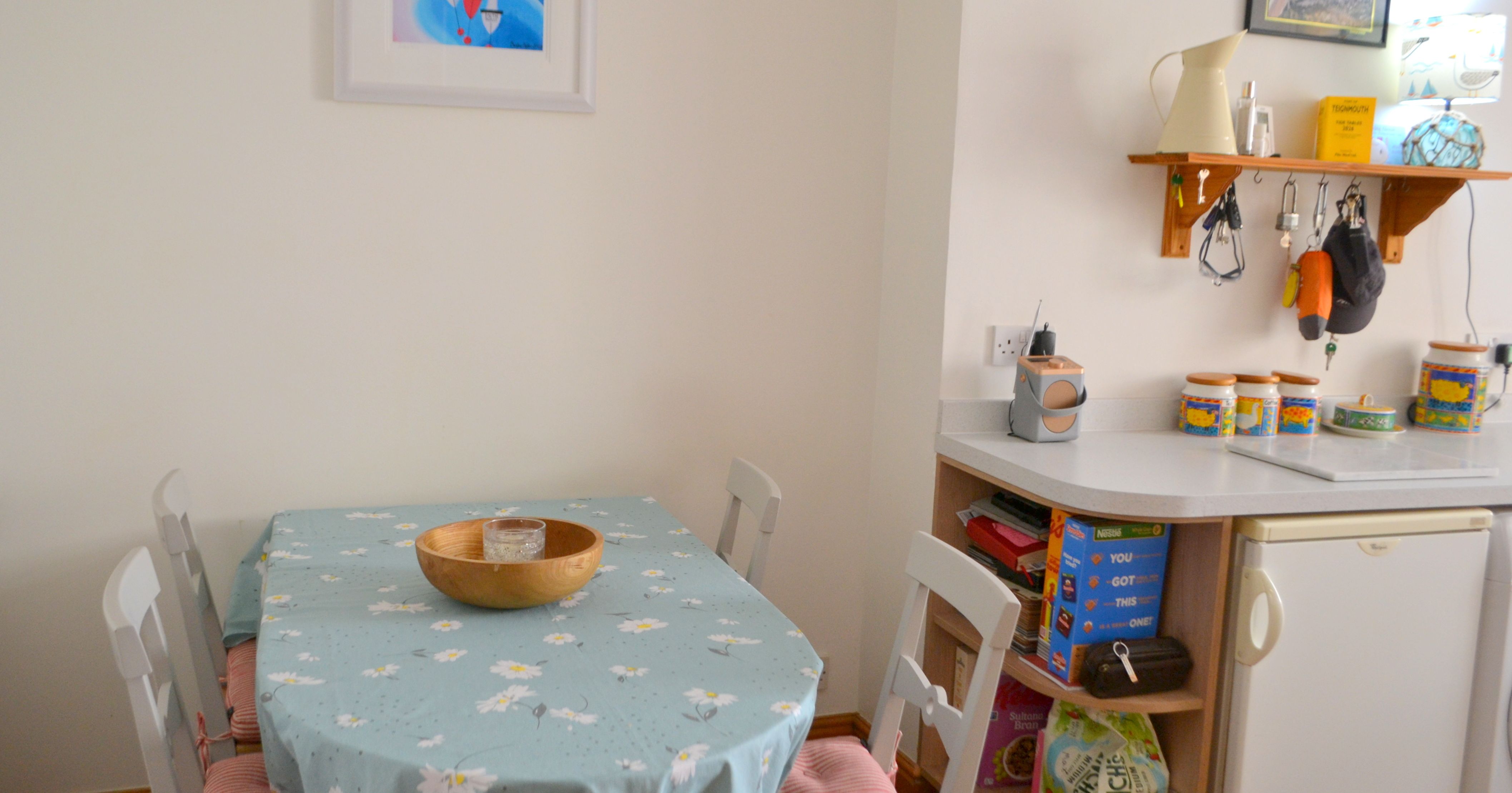 Small kitchen dining area with a table covered in a blue daisy tablecloth, four chairs, and a bowl as centerpiece, with a shelf and kitchen items in the background.
