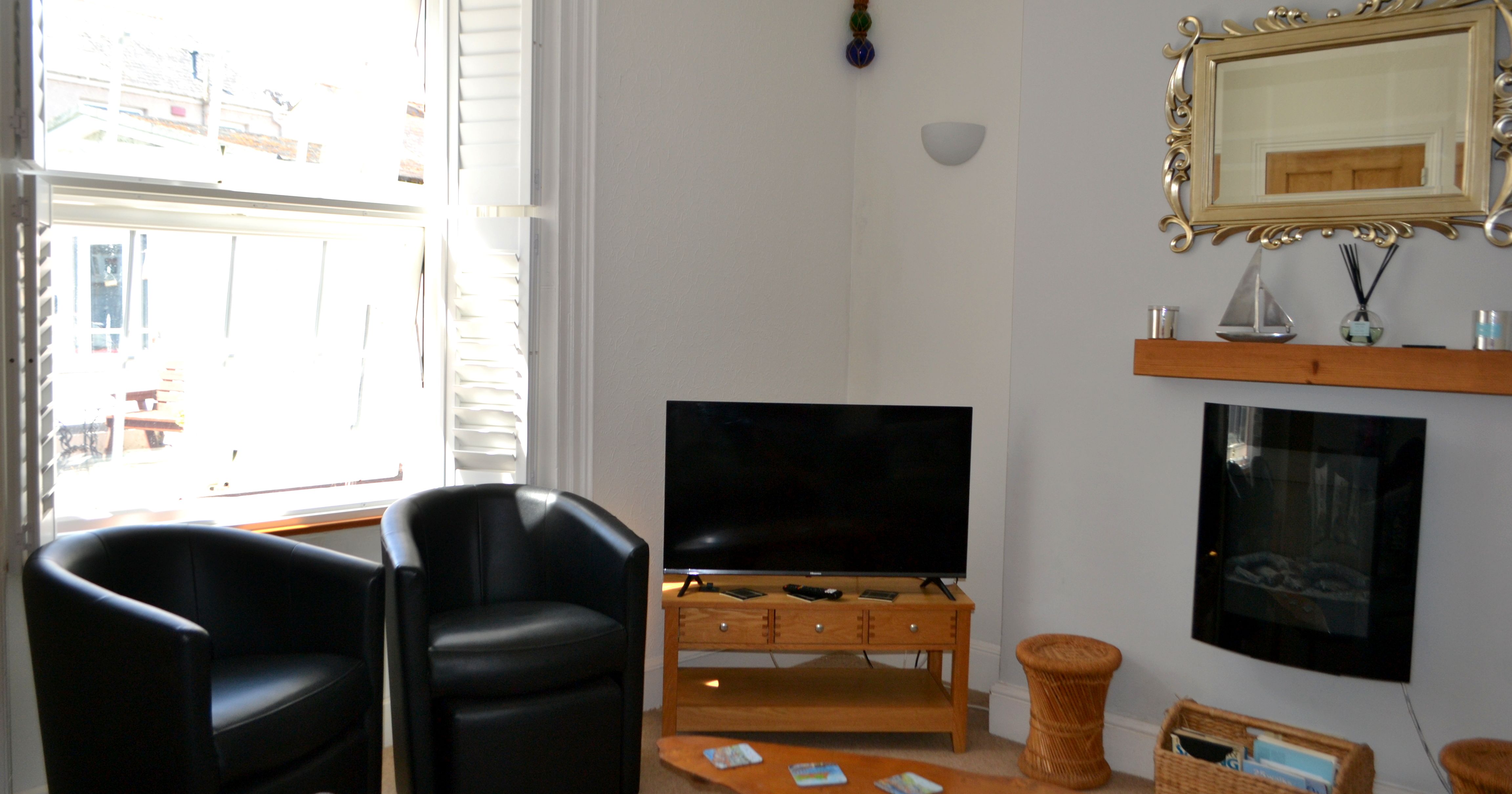 Modern living room with two black chairs, a TV on a wooden stand, a large window with white shutters, a decorative mirror, and a wooden coffee table.