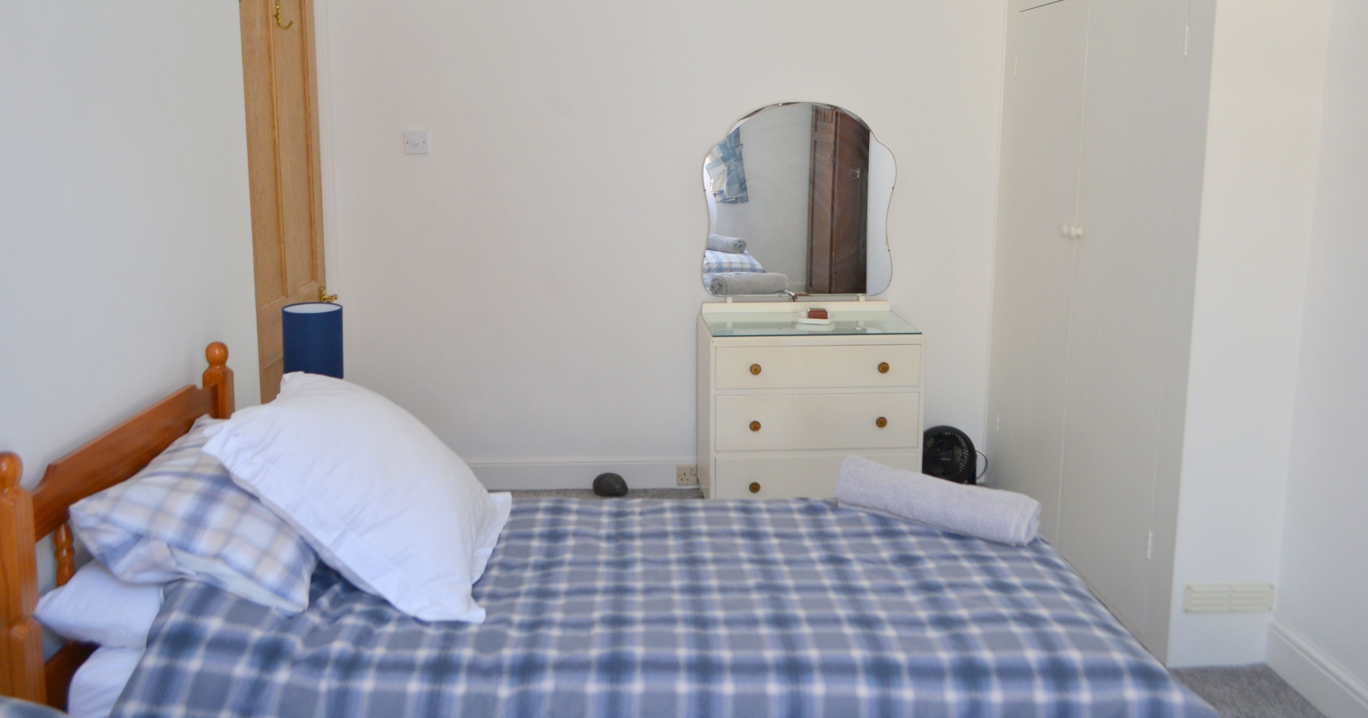 A simple twin bedroom with blue checkered bedding, a wooden bed frame, wardrobe, and a dresser with a mirror.
