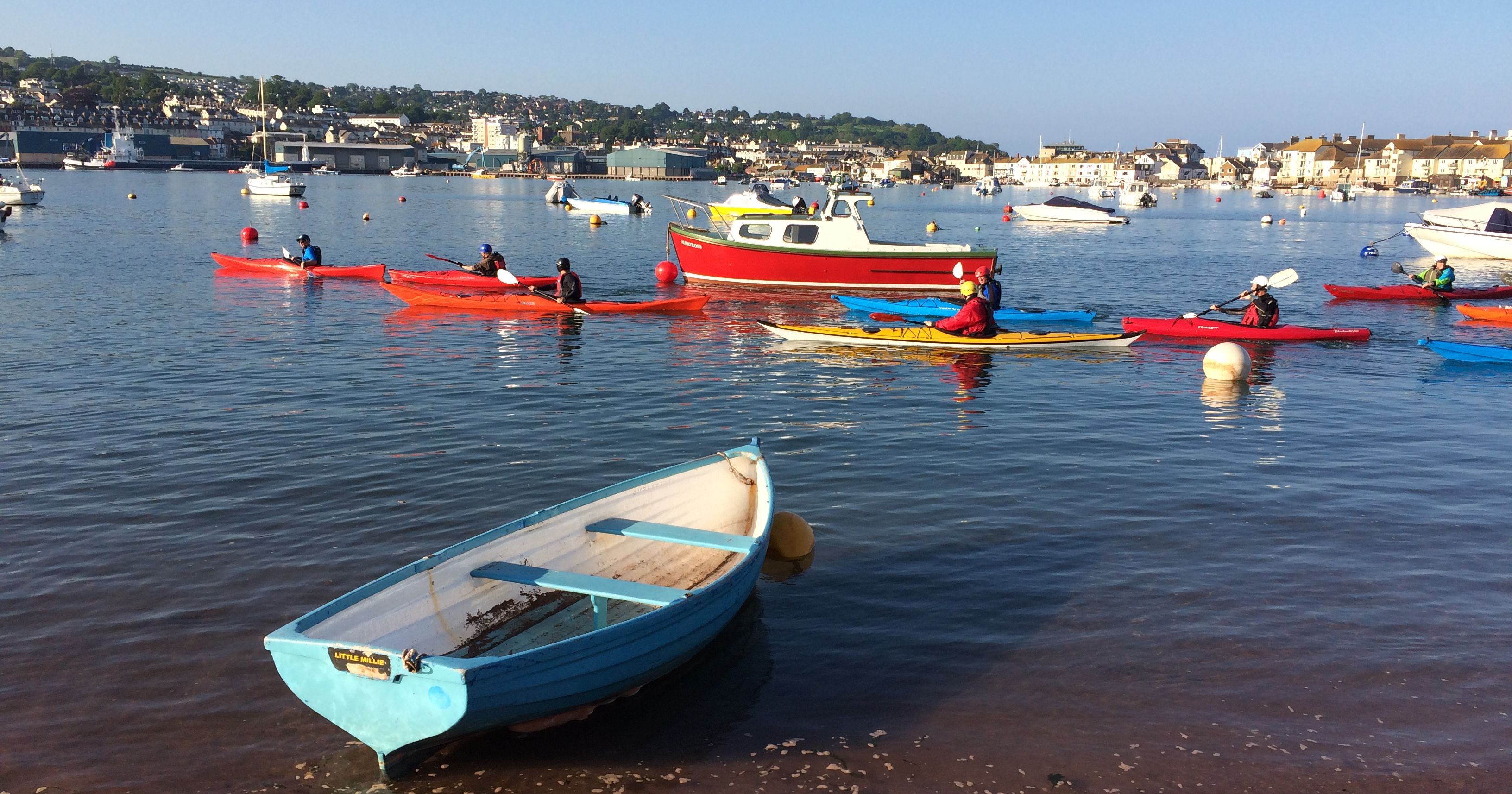 Colorful kayaks and boats floating in a harbor with people paddling on a sunny day.