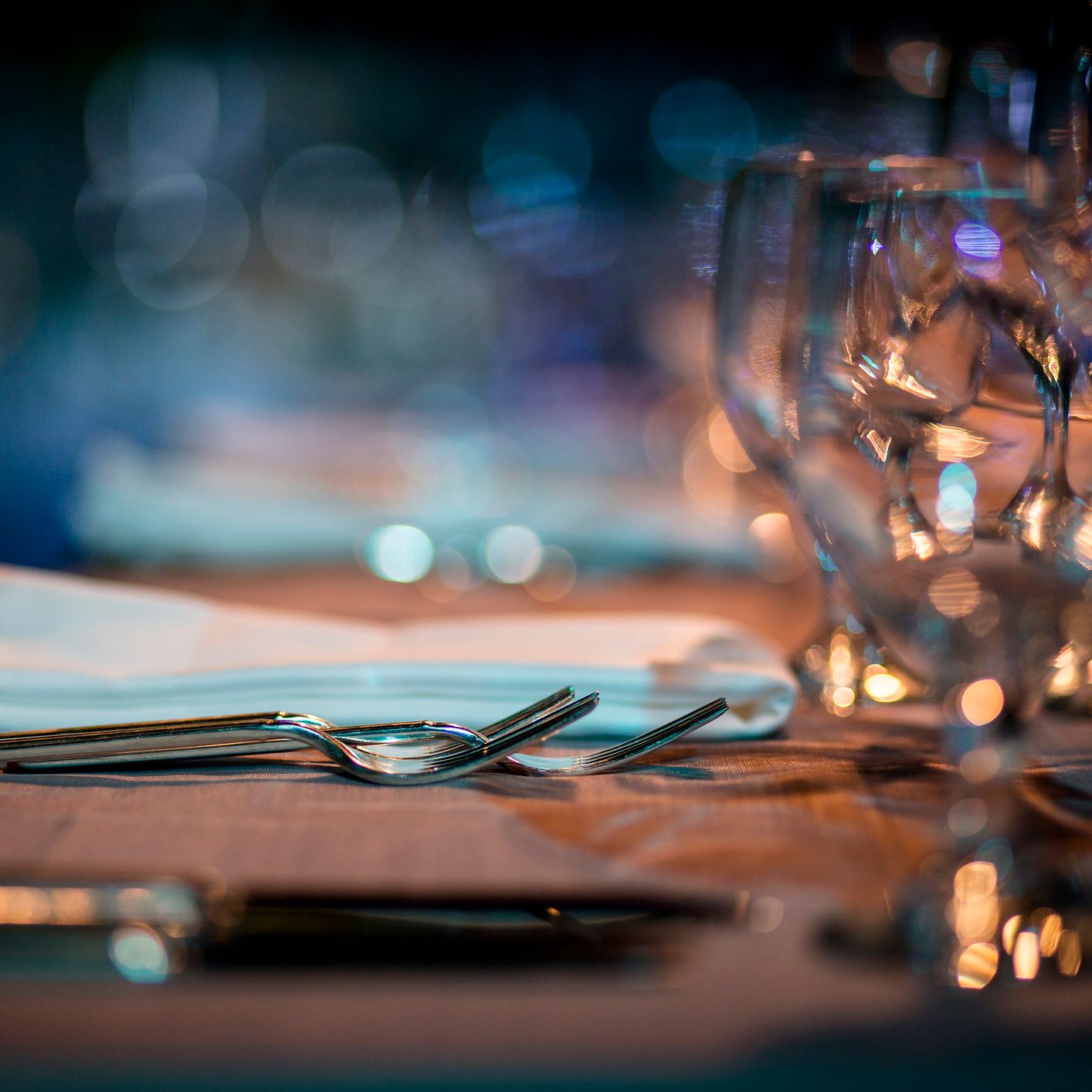 A close-up of an elegant dining table setting with polished cutlery, neatly folded napkins, and sparkling glassware, illuminated by warm ambient lighting.
