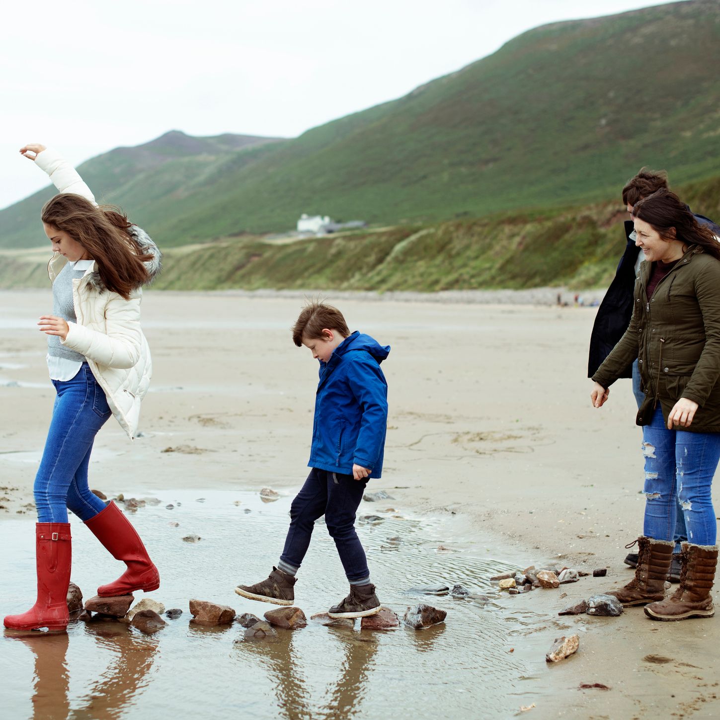 A family enjoys a day at the beach, with children in wellies stepping on rocks across a shallow stream, while parents watch and smile against a scenic hillside backdrop.