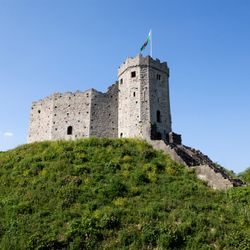A historic stone castle with a round tower stands atop a green hill, with a Welsh flag flying against a clear blue sky, showcasing medieval architecture.