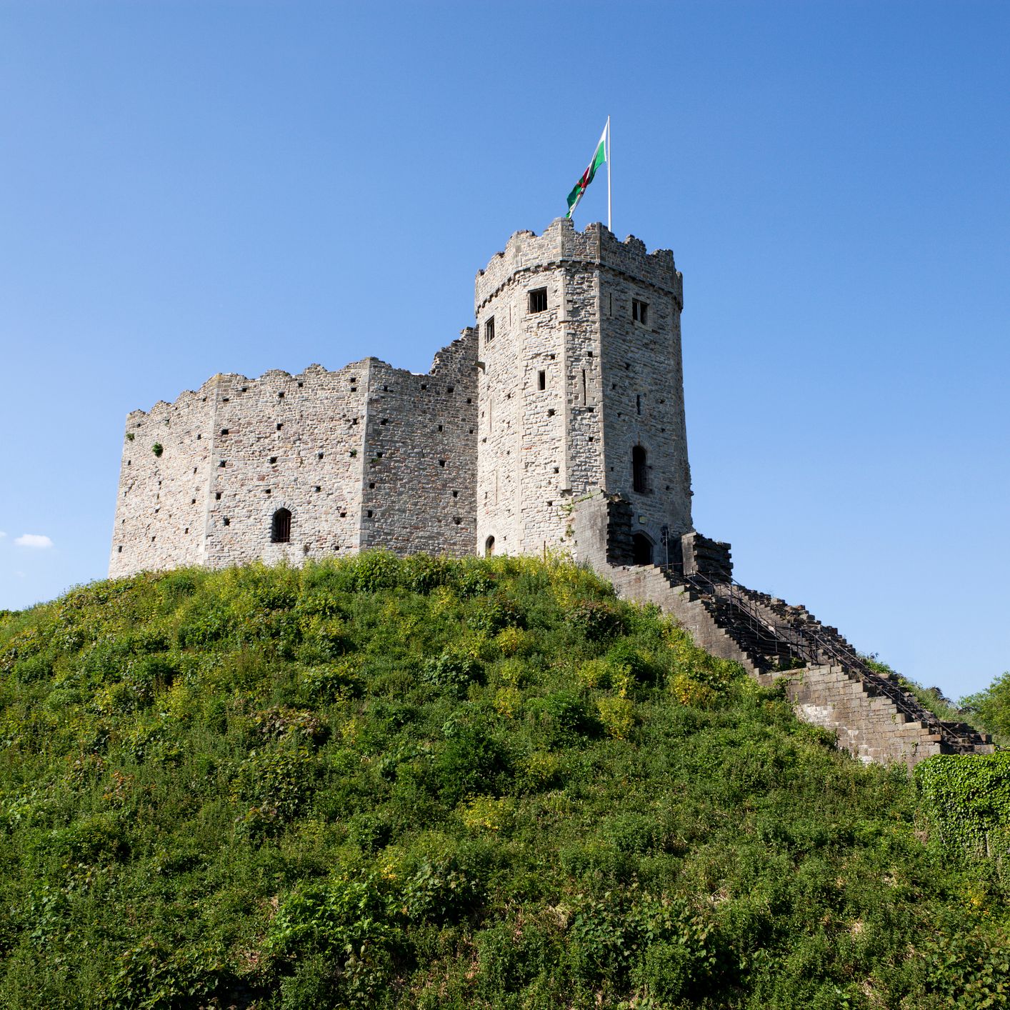 A historic stone castle with a round tower stands atop a green hill, with a Welsh flag flying against a clear blue sky, showcasing medieval architecture.