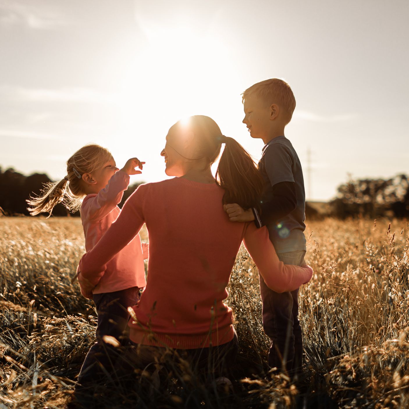 A mother and her two children enjoy a sunny afternoon in a field, laughing and playing together as the golden light of sunset casts a warm glow over the scene.