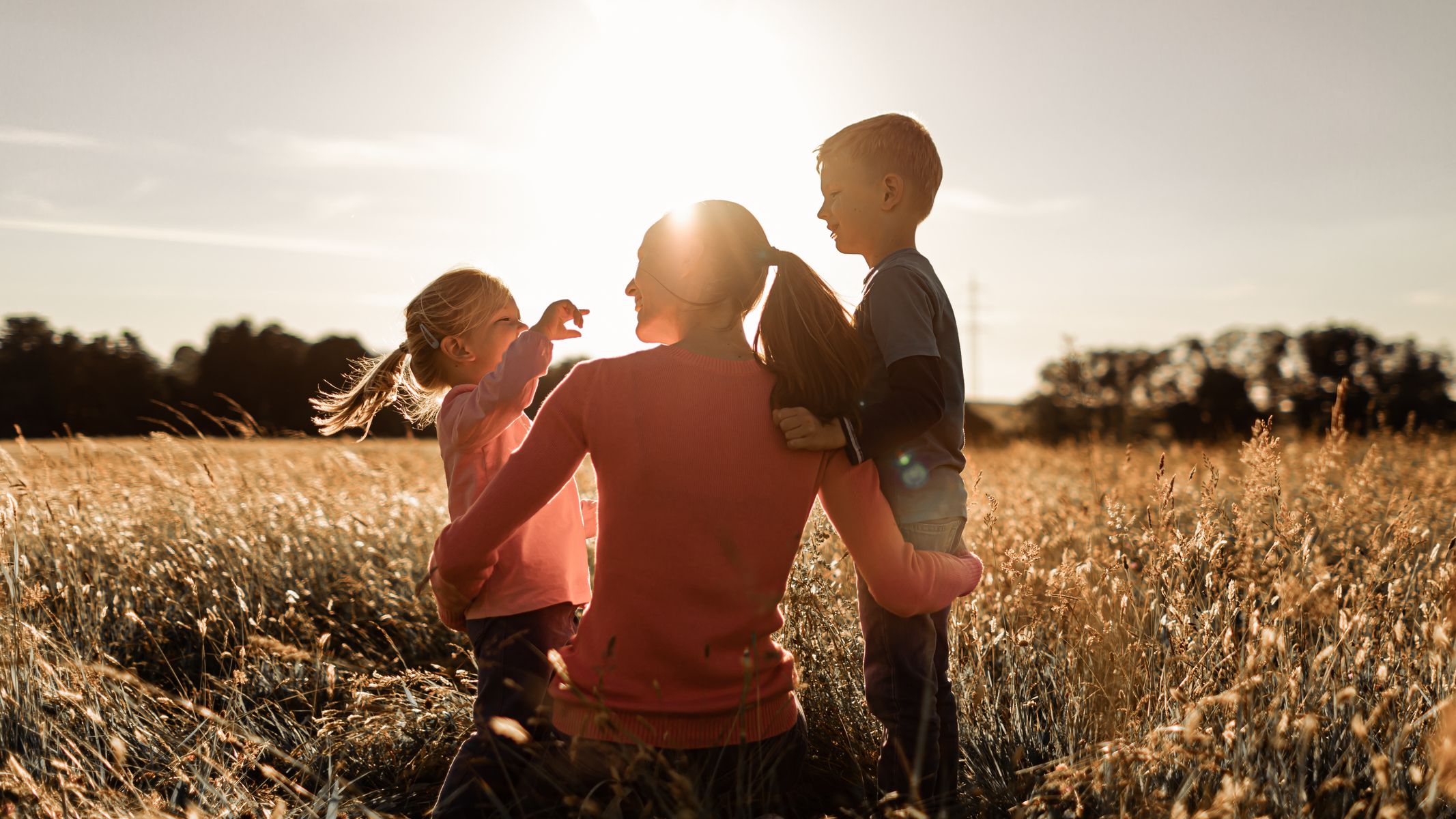A mother and her two children enjoy a sunny afternoon in a field, laughing and playing together as the golden light of sunset casts a warm glow over the scene.