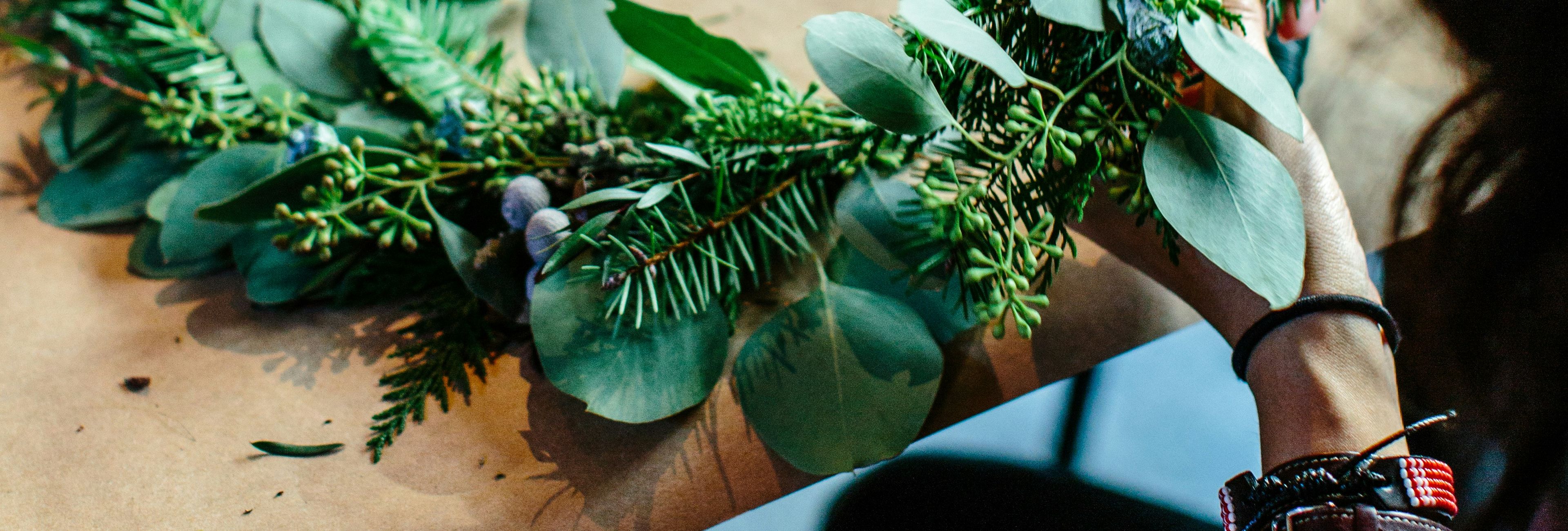Person making a wreath with green foliage and eucalyptus leaves on a table