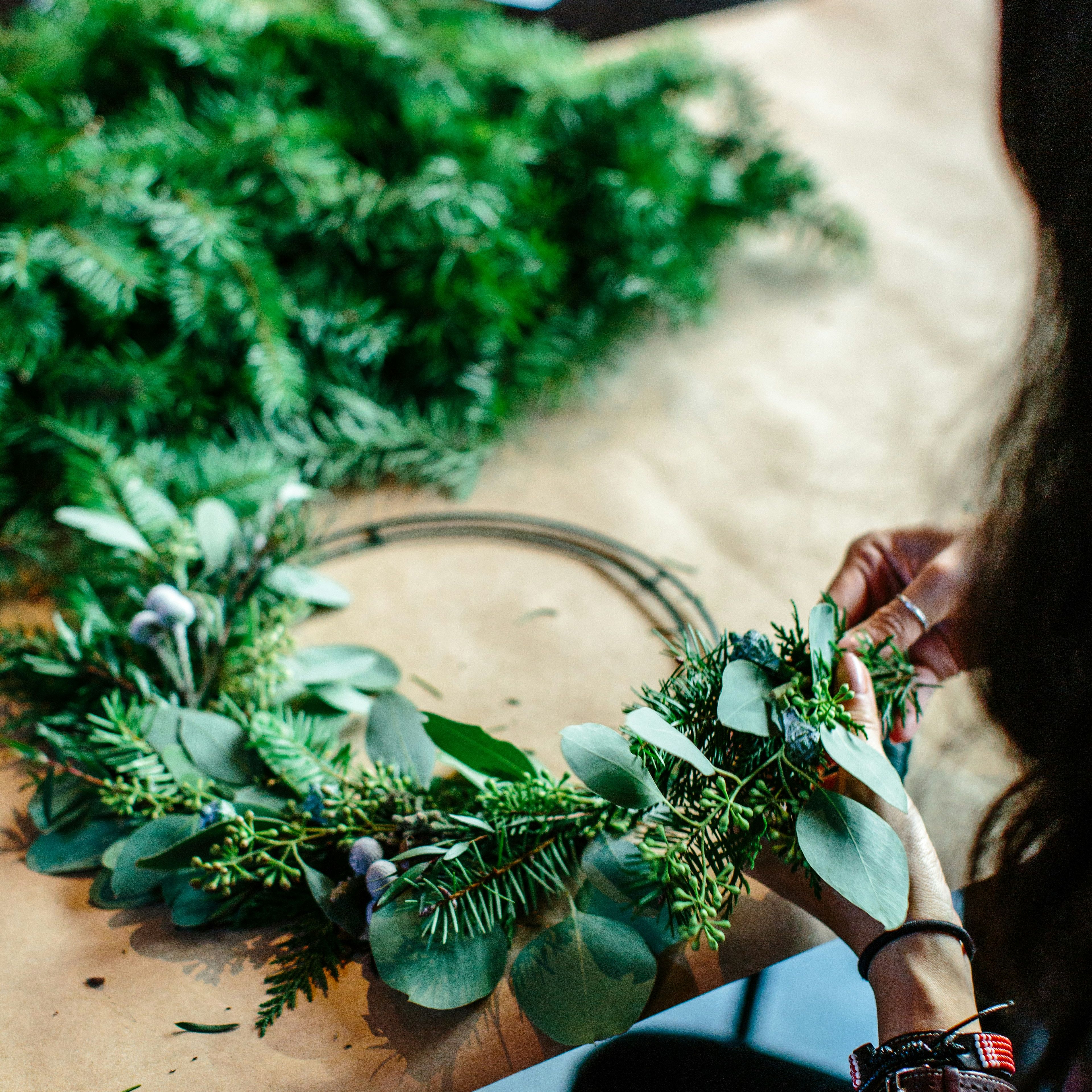 Person making a wreath with green foliage and eucalyptus leaves on a table