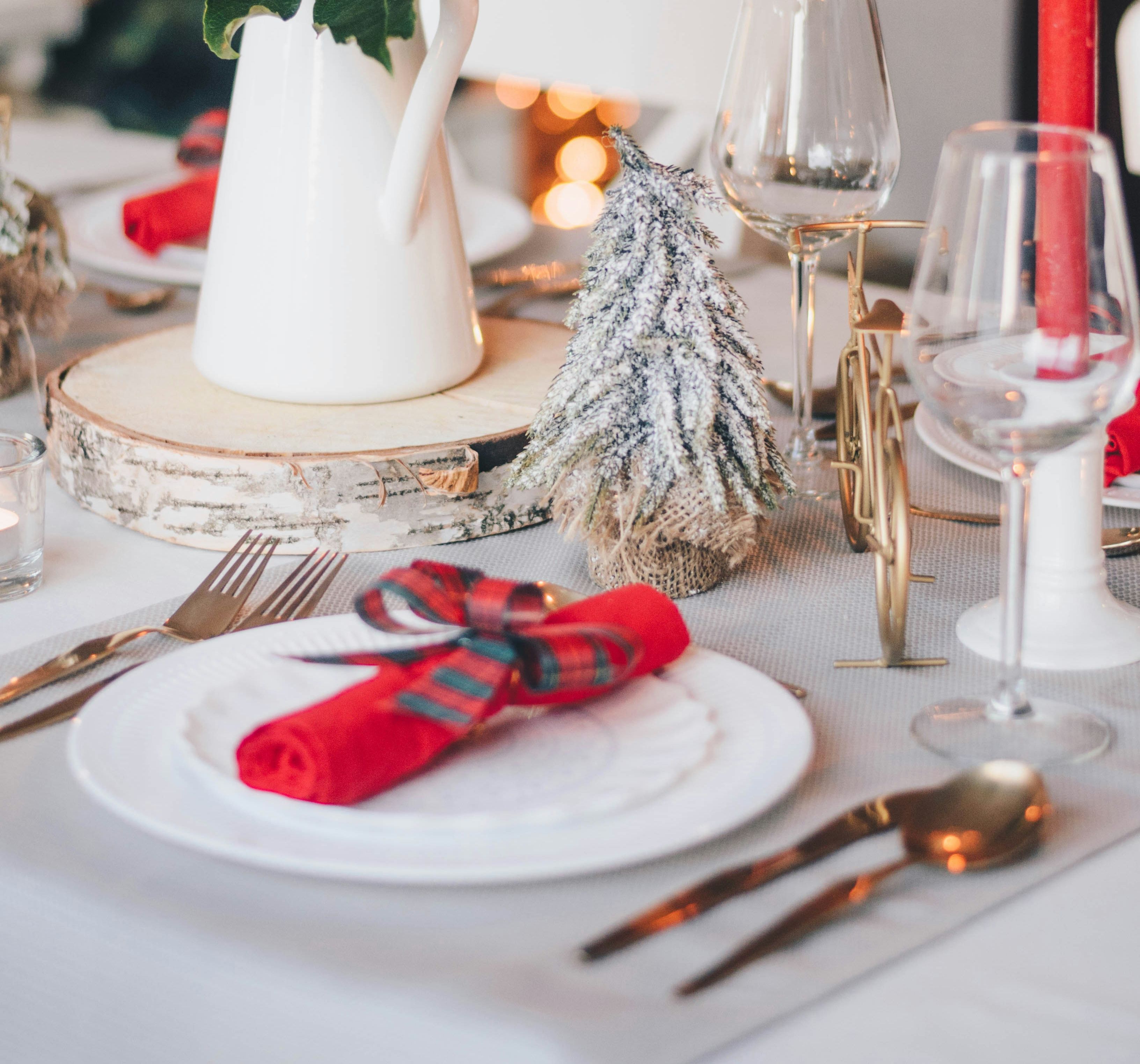 Festive holiday table setting with a white pitcher of red flowers, a mini Christmas tree, red napkins, and lit candles.