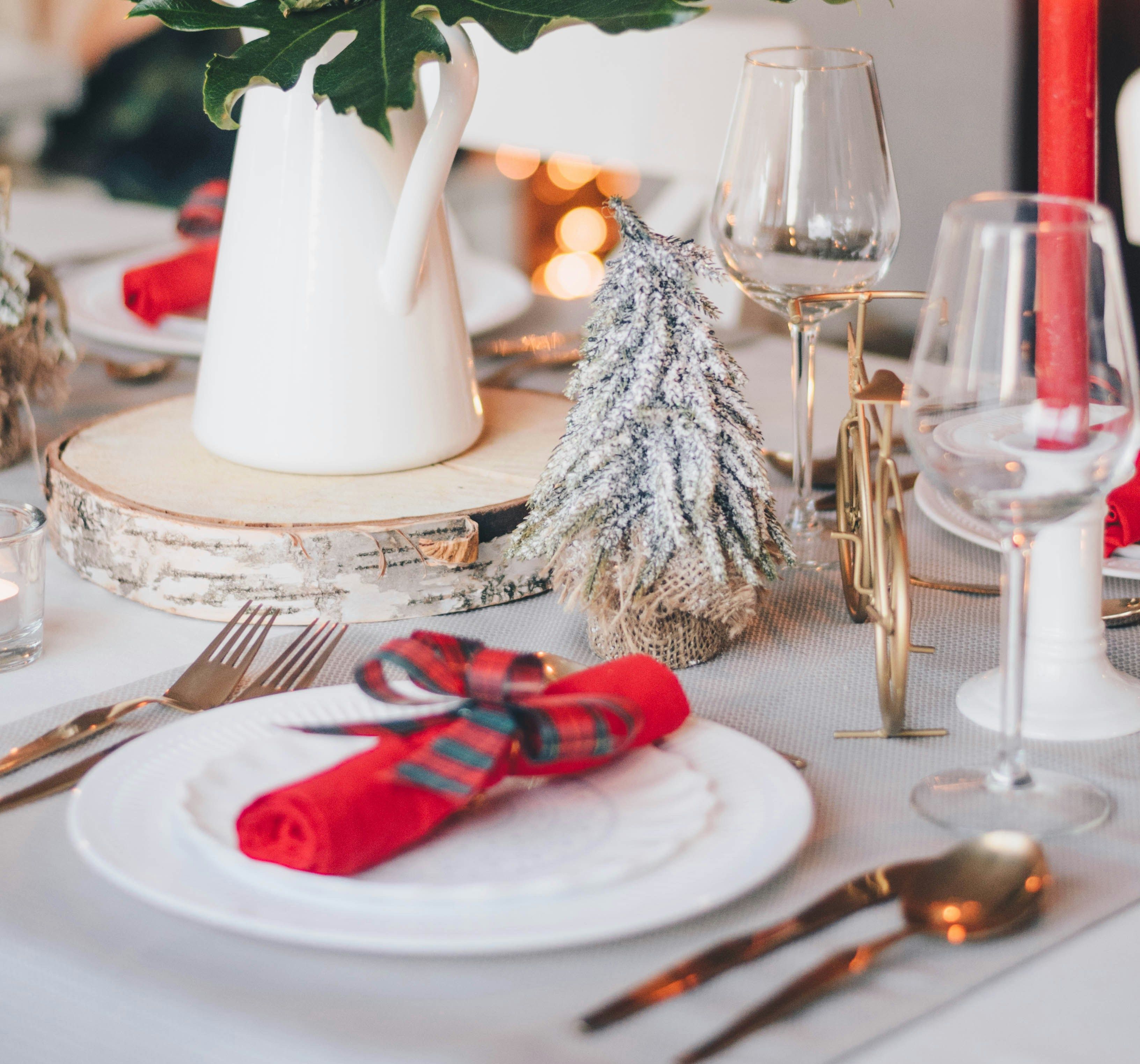 Festive holiday table setting with a white pitcher of red flowers, a mini Christmas tree, red napkins, and lit candles.