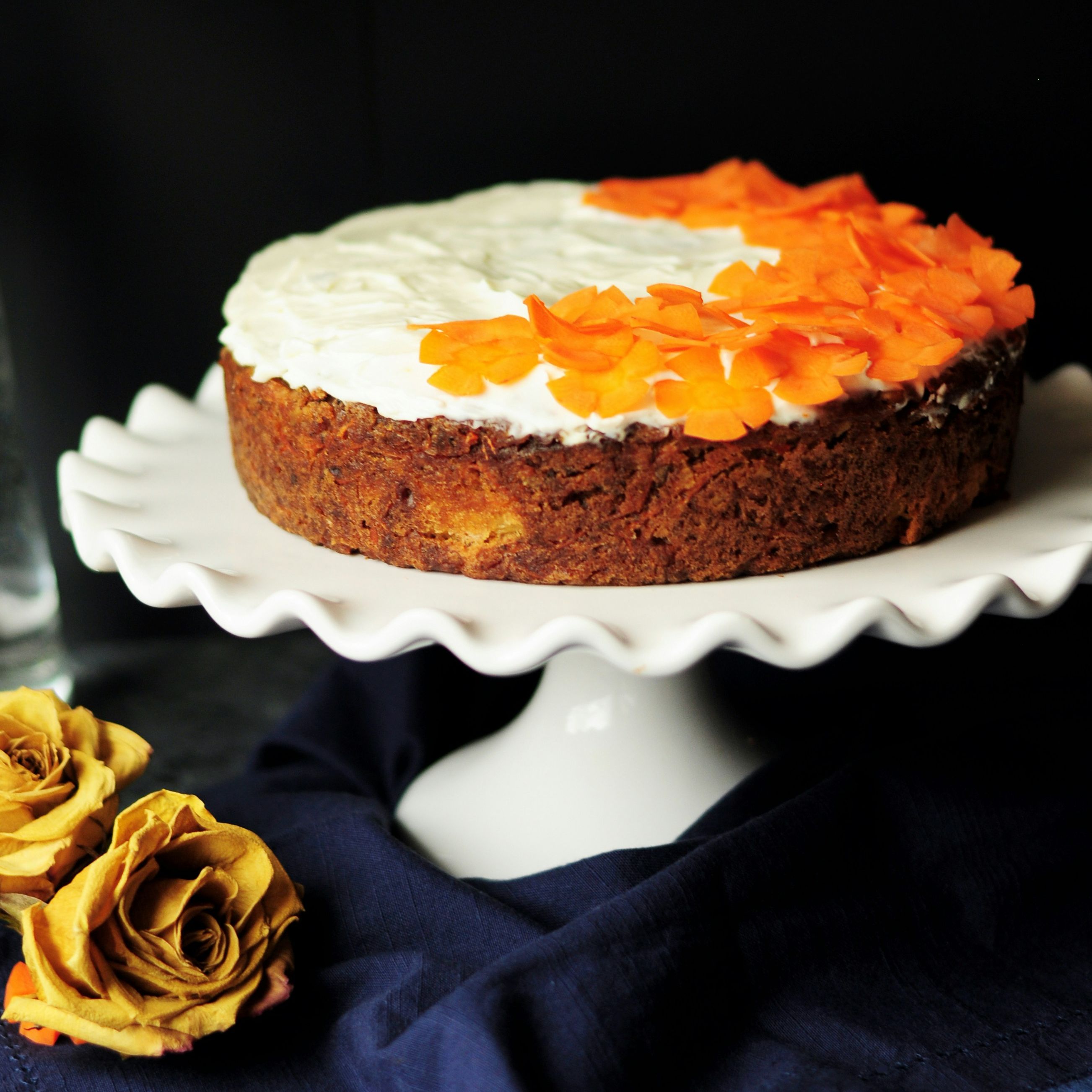 Carrot cake with white frosting and orange flower-shaped decorations on top, displayed on a white cake stand