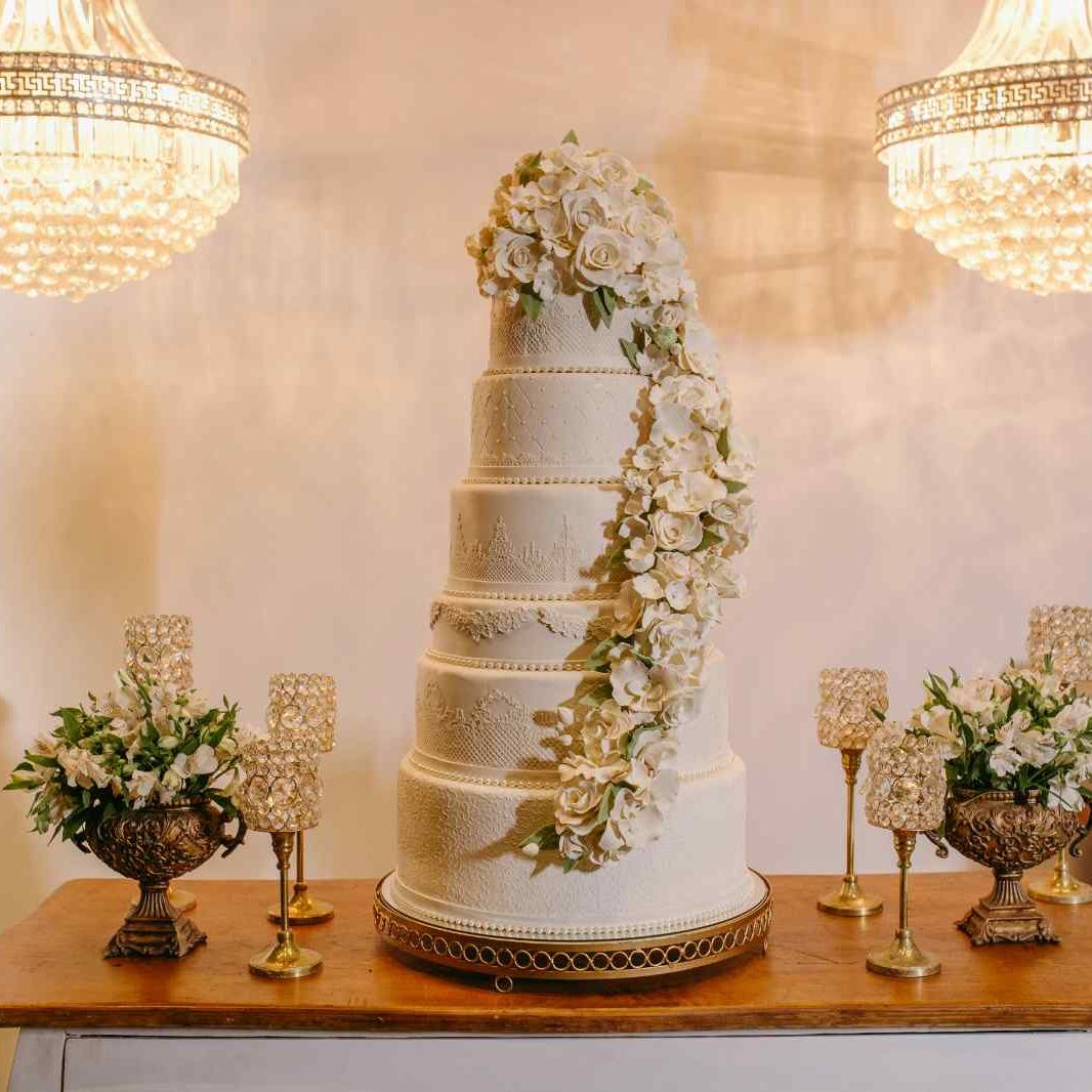 Elegant multi-tiered white wedding cake adorned with cascading white flowers, displayed on a wooden table with decorative flowers and golden candle holders.