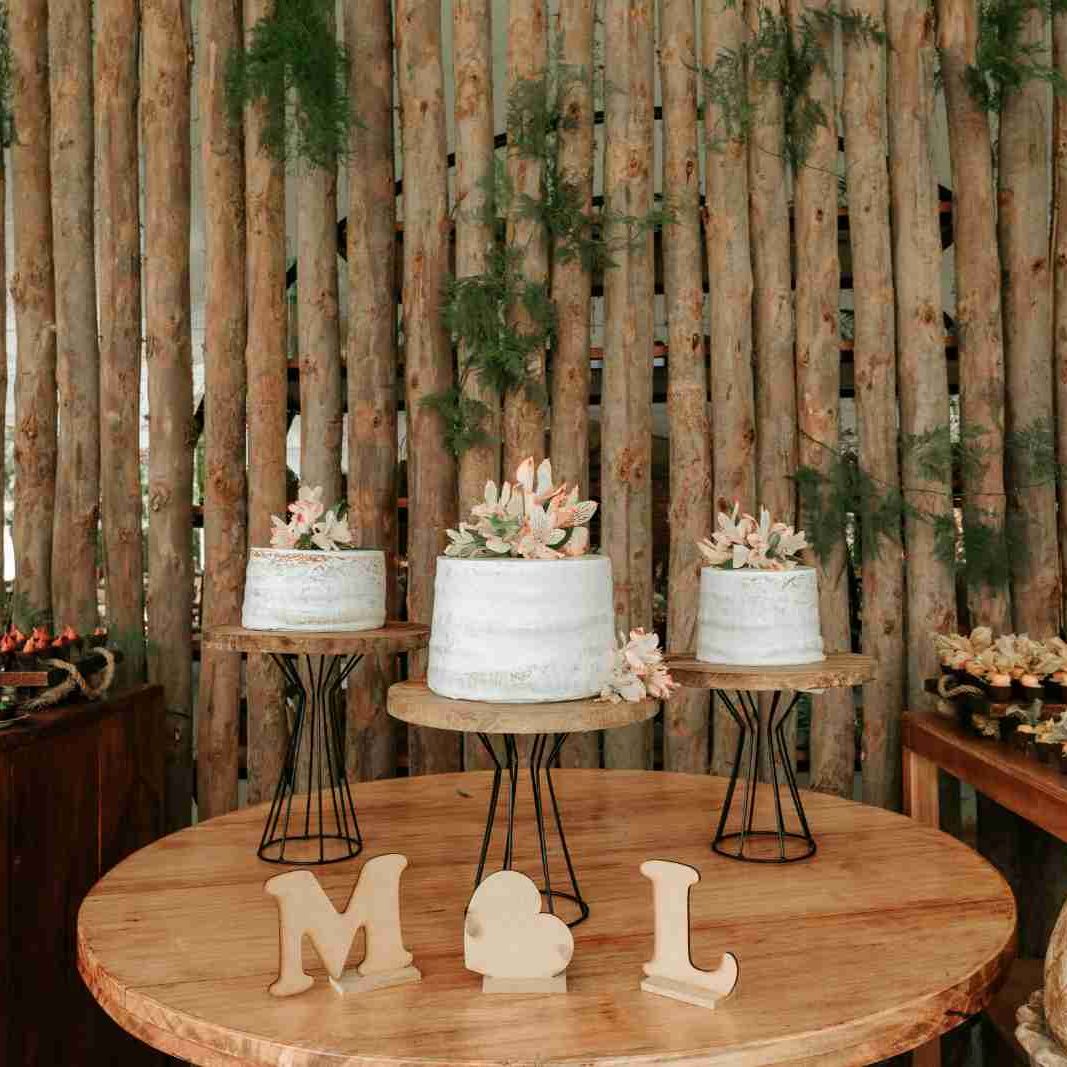 Three white cakes with floral decorations on wooden stands, rustic background with wooden logs and greenery.