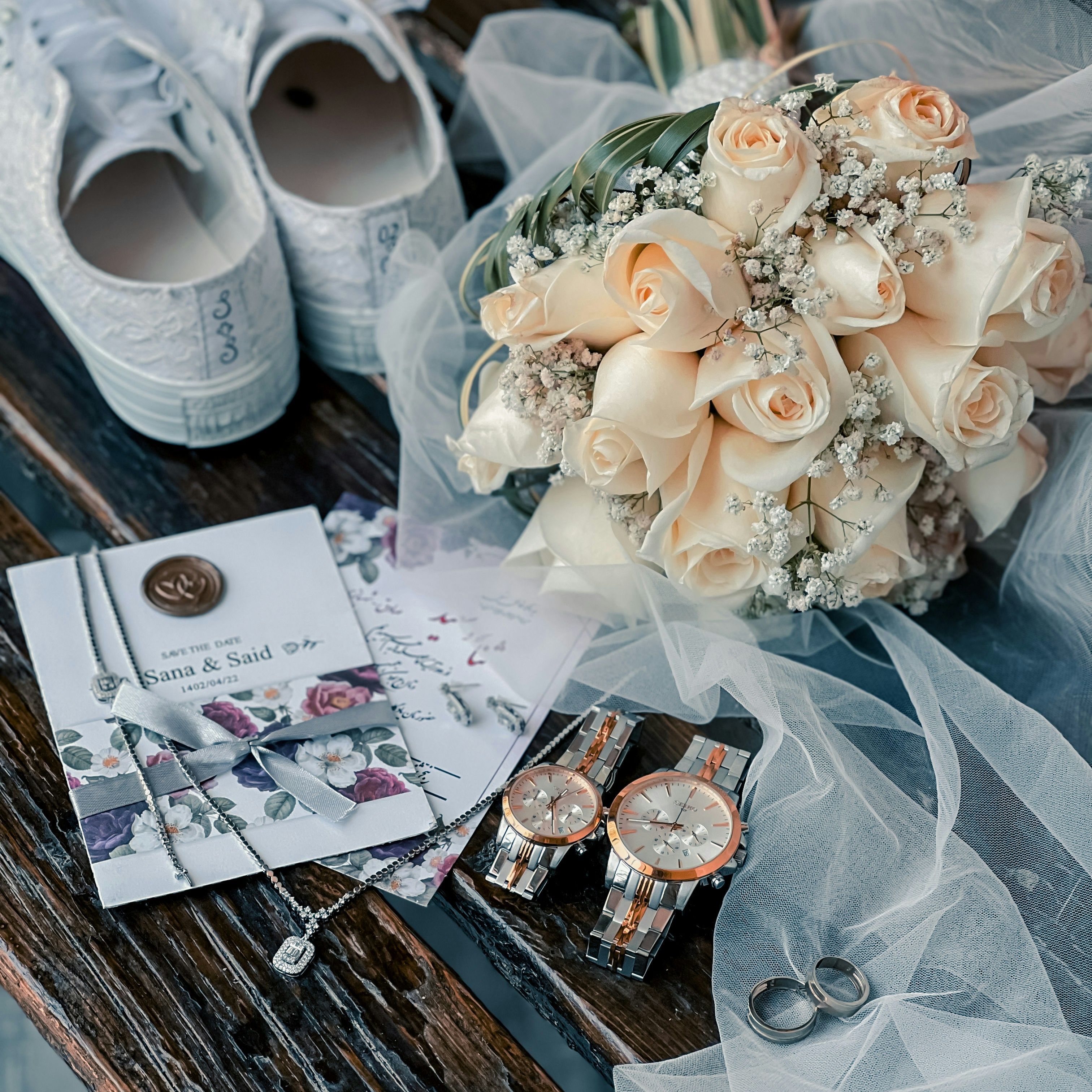 Wedding details on a wooden bench with bridal shoes, bouquet of pale roses, invitation, watches and rings