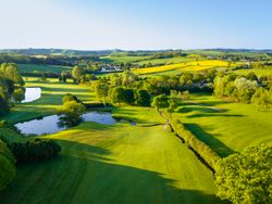 Aerial view of a golf course surrounded by green fields, trees, ponds, and rolling countryside.
