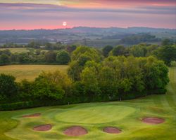 Golf course green with sand bunkers at sunrise, surrounded by lush greenery and rolling hills.