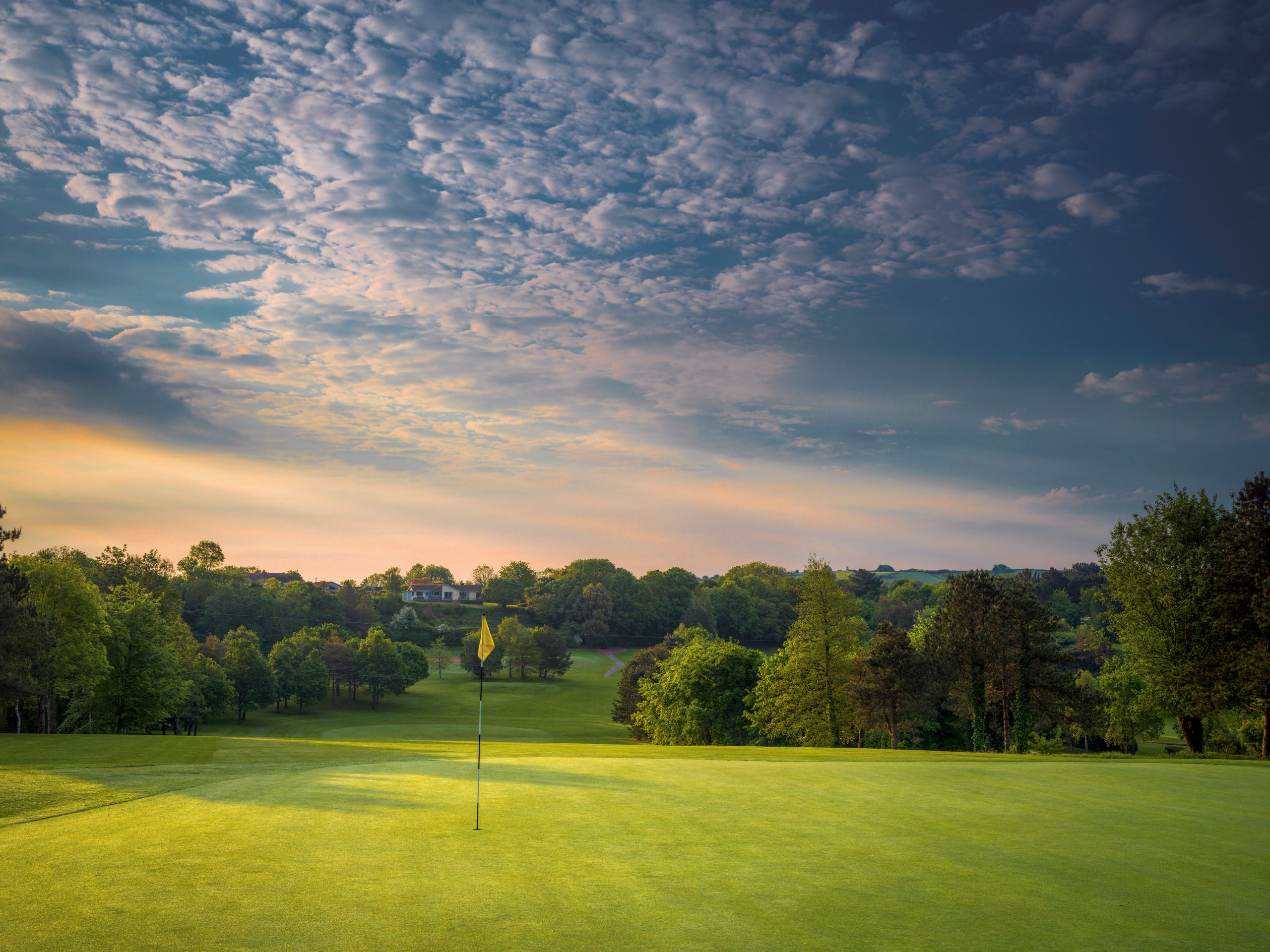 A golf course green with a flagstick, surrounded by trees under a dramatic sky at sunset.