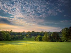 A golf course green with a flagstick, surrounded by trees under a dramatic sky at sunset.
