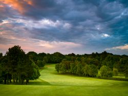 A lush green golf course fairway with trees and a dramatic sunset sky overhead.