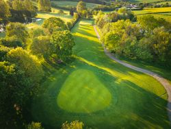 Aerial view of a golf course green surrounded by trees and a winding path during golden hour.