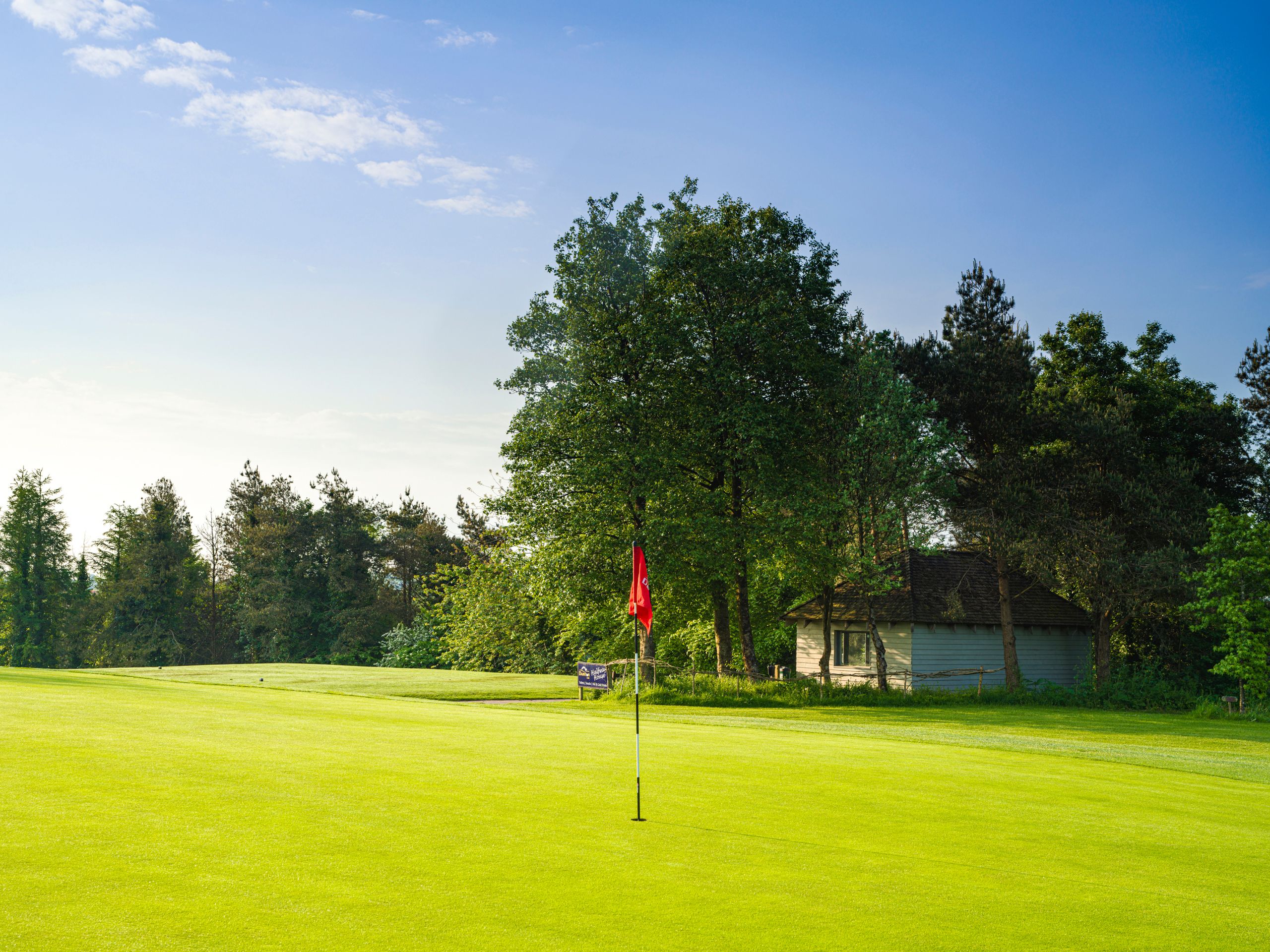 Golf course green with a red flag and a small building among trees in the background
