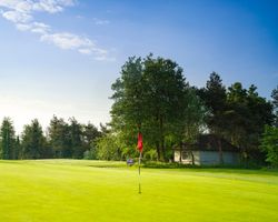 Golf course green with a red flag and a small building among trees in the background