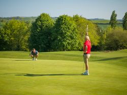 Two golfers on a green, one crouching and aiming a putt, the other standing and holding the flagstick.
