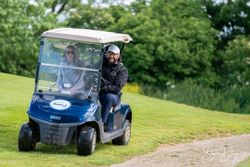 Two people riding a golf cart on a grassy course, one smiling and driving, the other sitting beside.