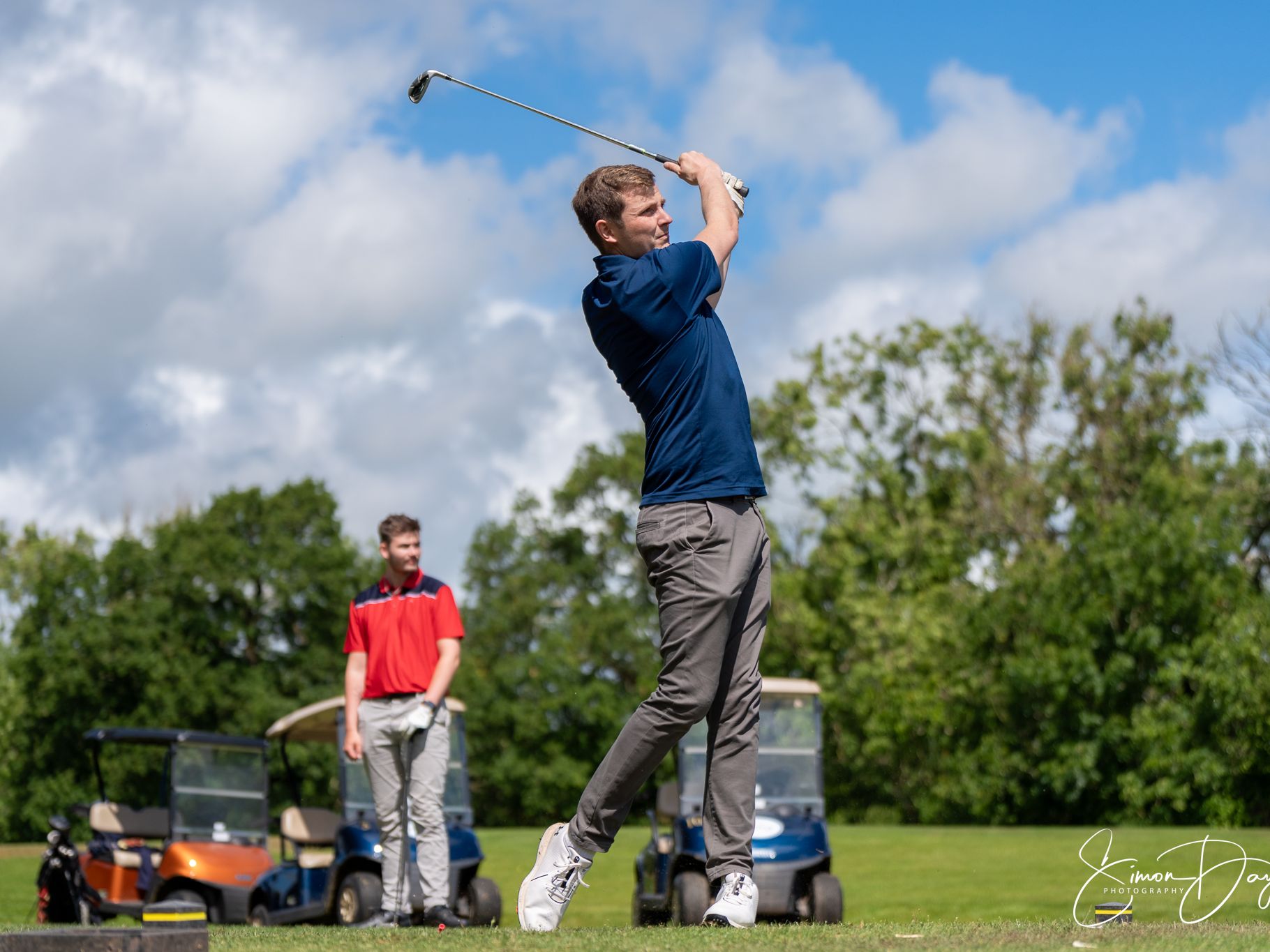 Man taking a golf swing with another person watching, golf carts in the background
