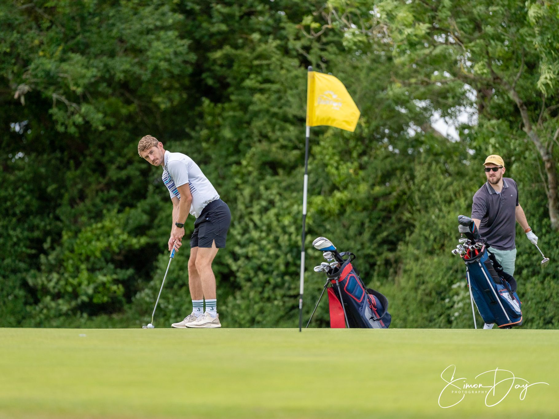 Two men on a golf course, one putting while the other stands by their golf bags near a yellow flag.