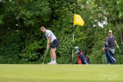 Two men on a golf course, one putting while the other stands by their golf bags near a yellow flag.